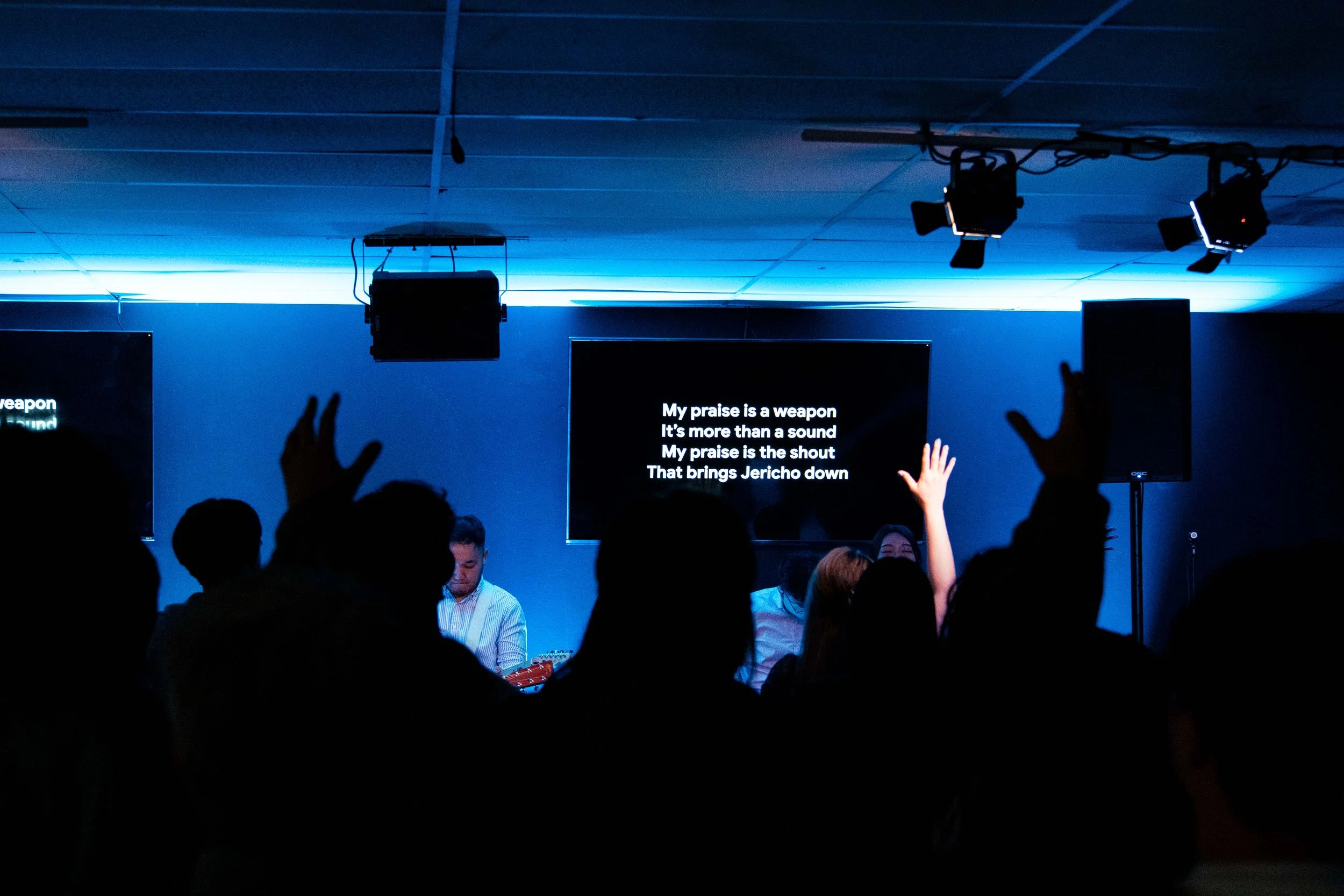 A scene inside a darkened room with a blue-lit ceiling, showing a group of people with some raising their hands, and a screen displaying song lyrics.