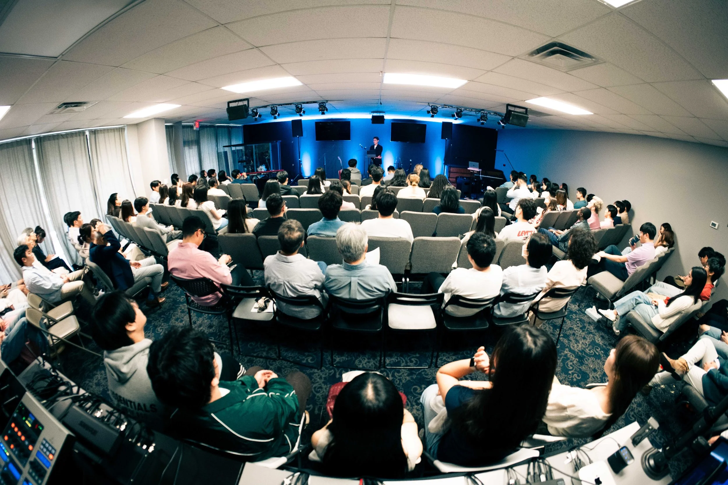 An audience of diverse people seated in an auditorium, attentively listening to a speaker on stage, with blue lighting and three large screens behind the speaker.