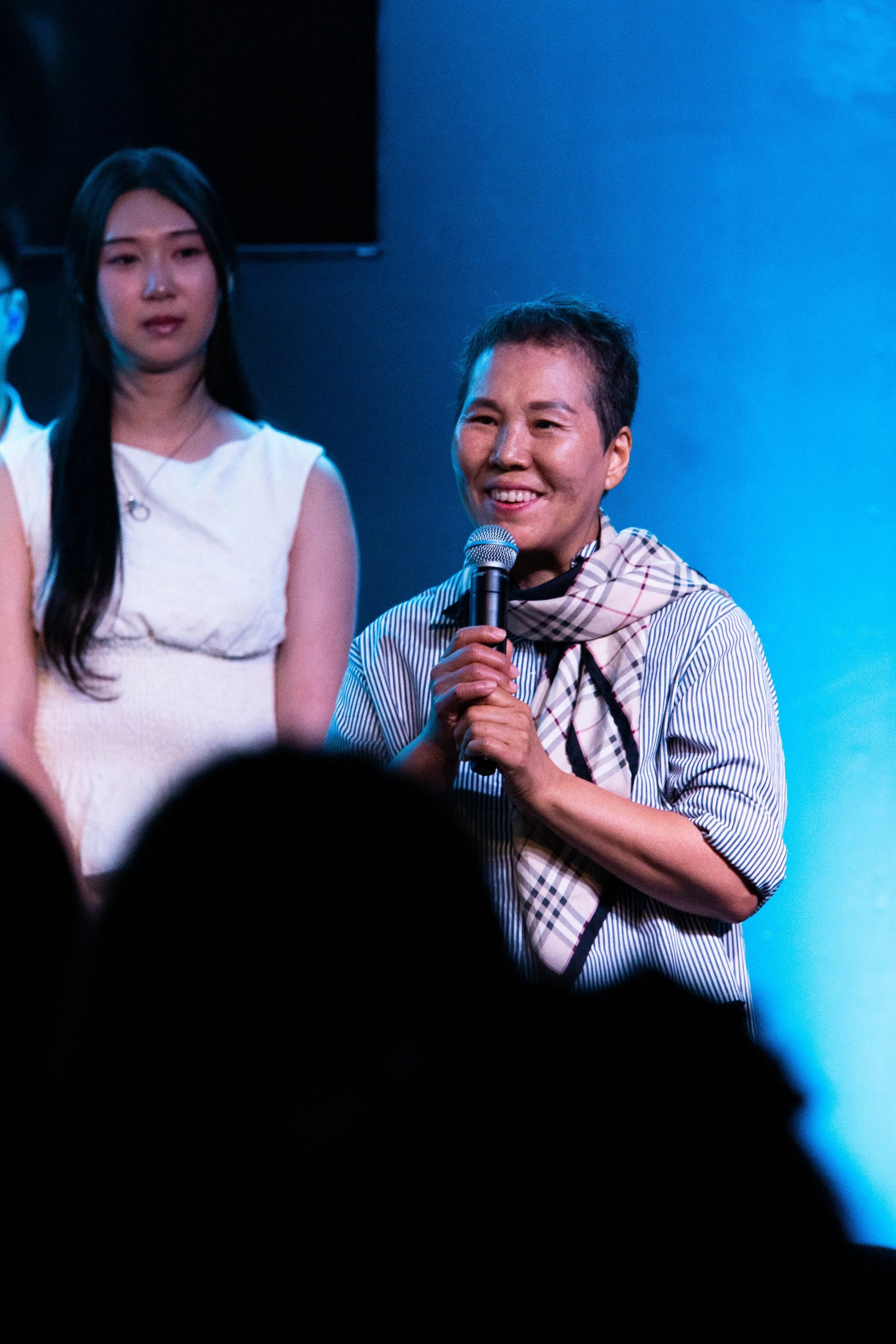 A woman with short dark hair smiling while holding a microphone on stage, standing next to a woman with long dark hair wearing a white dress, against a blue background.