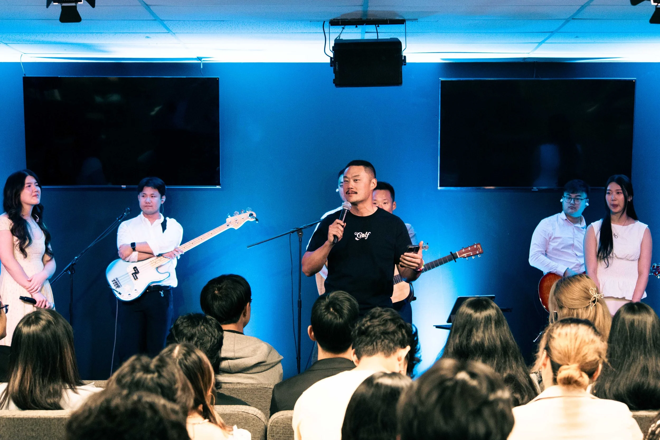 A man with a microphone and a phone stands in front of a group of people on a stage. Several people stand on the stage with musical instruments, including a bass guitar. The audience is seated, and the stage has a blue background with large TV screens.