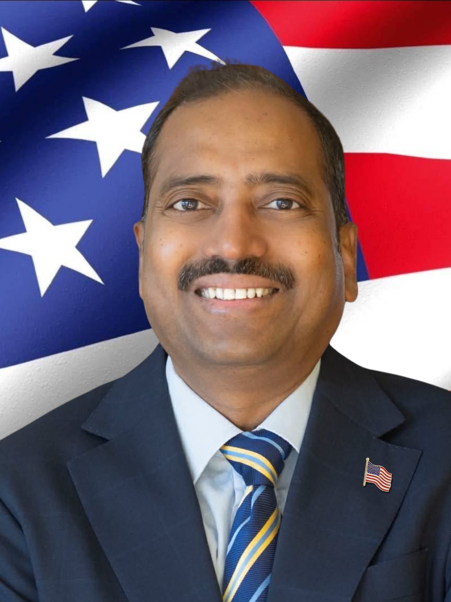 A man in a navy blue suit, light blue shirt, and a navy blue and yellow striped tie, smiling, with a small American flag pin on his lapel, in front of a large American flag.