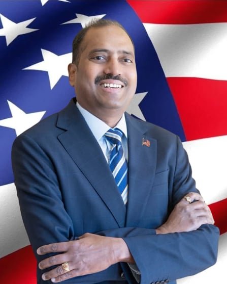 A man in a navy blue suit and striped tie standing in front of an American flag background smiling