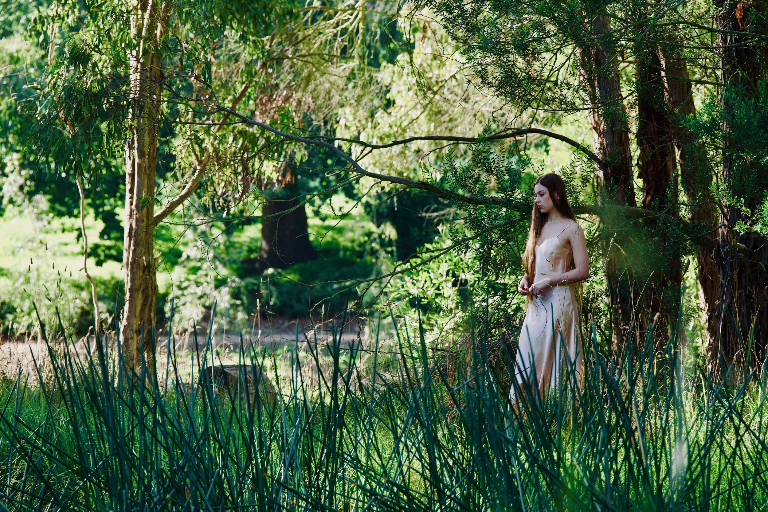 A young woman in a satin dress standing in a lush green forest among trees and tall grass.