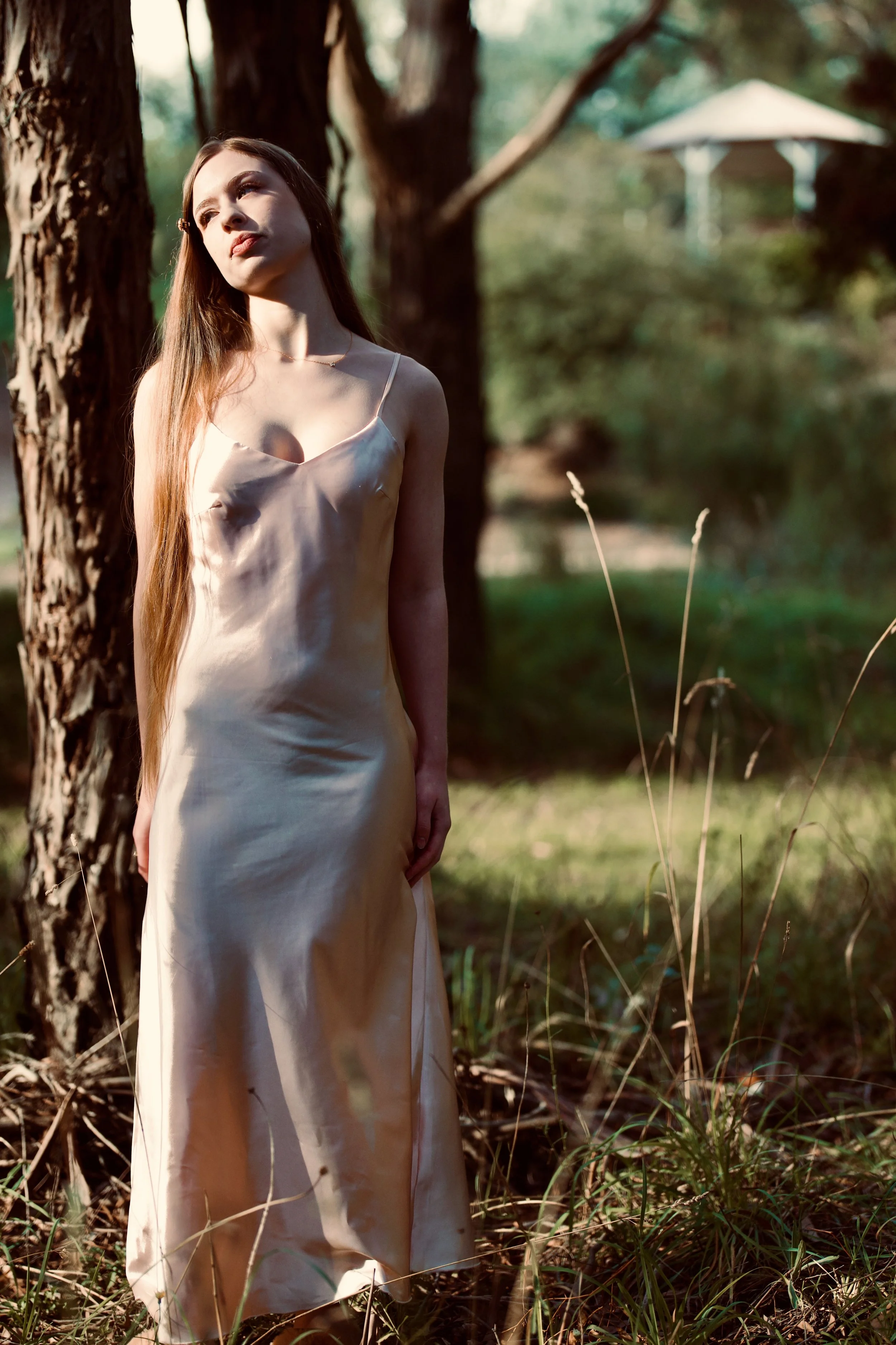 A woman with long hair wearing a satin dress standing outdoors near trees and grass, with sunlight shining on her face, and a blurred background including a white structure.