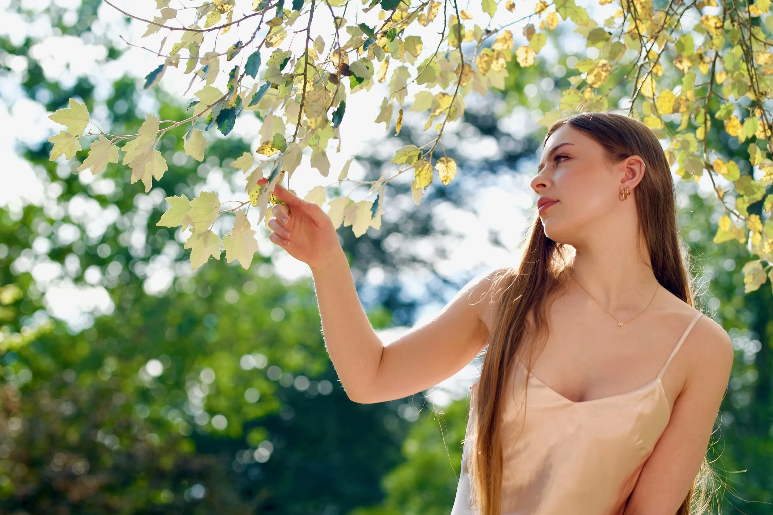 A young woman with long brown hair and gold earrings touching the leaves of a tree in a park on a sunny day.