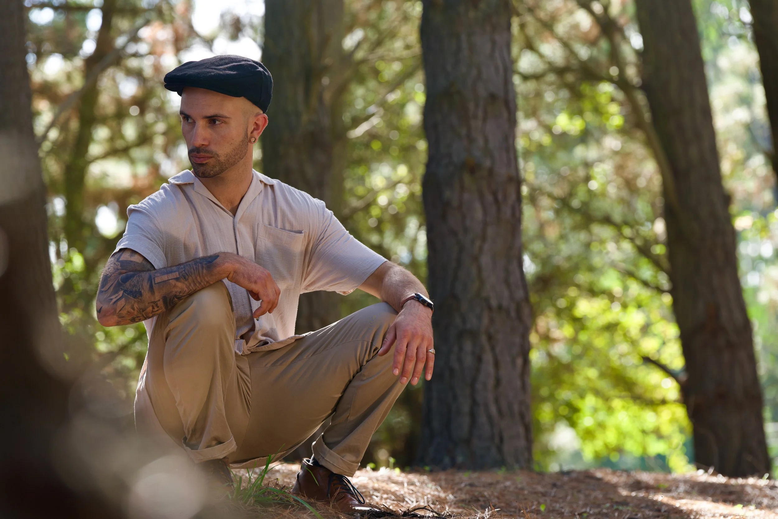 A man with tattoos on his arm wearing a black beret, beige shirt, and beige pants, sitting on the ground in a wooded area with tall trees and green foliage in the background.