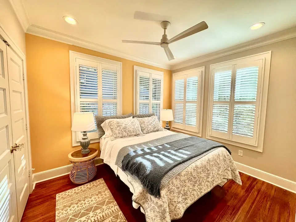 Cozy bedroom with a bed, lamps on side tables, large windows with white shutters, and hardwood flooring.