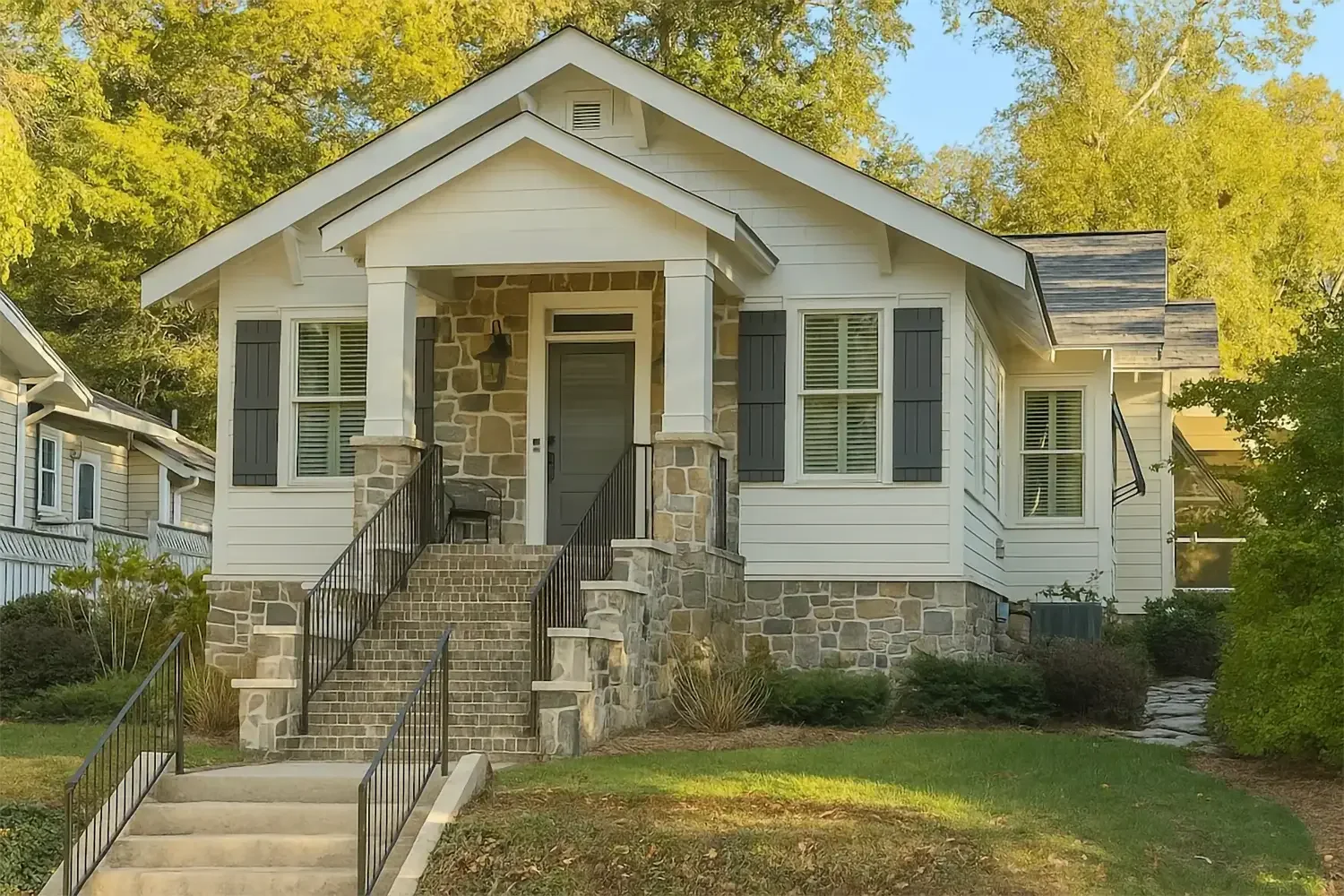 Front view of a house with white siding, a stone porch and steps, black railings, and dark shutters, surrounded by green trees and lawn.