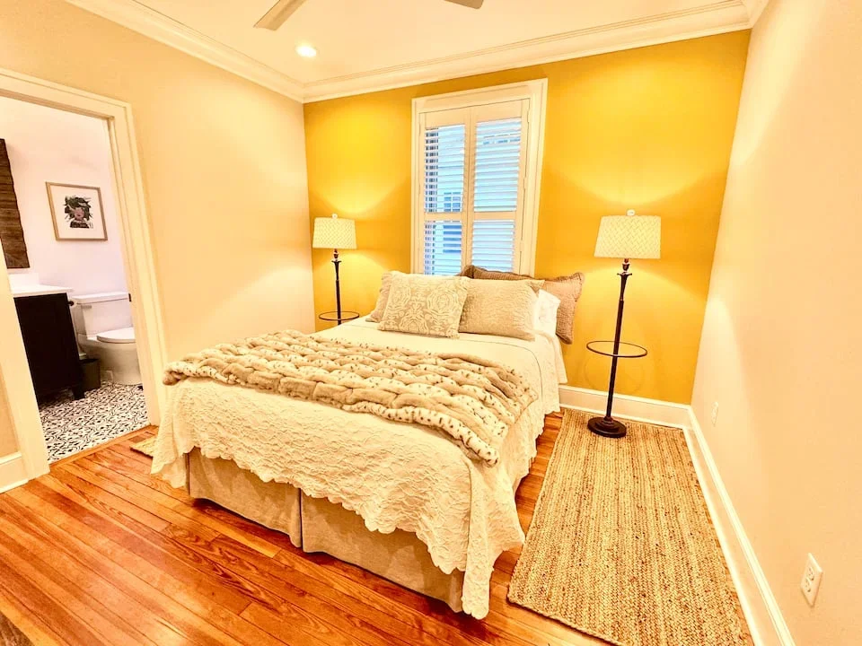 A cozy bedroom with a bed covered in white and beige bedding and decorative pillows. Two floor lamps provide warm lighting on either side of the bed. There is a window with closed blinds behind the bed. To the left, an open door shows a bathroom with patterned tile flooring.
