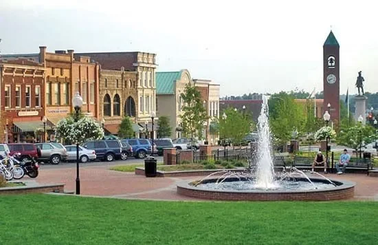 Town square with a fountain, historic buildings, parked cars, and a clock tower in the background.