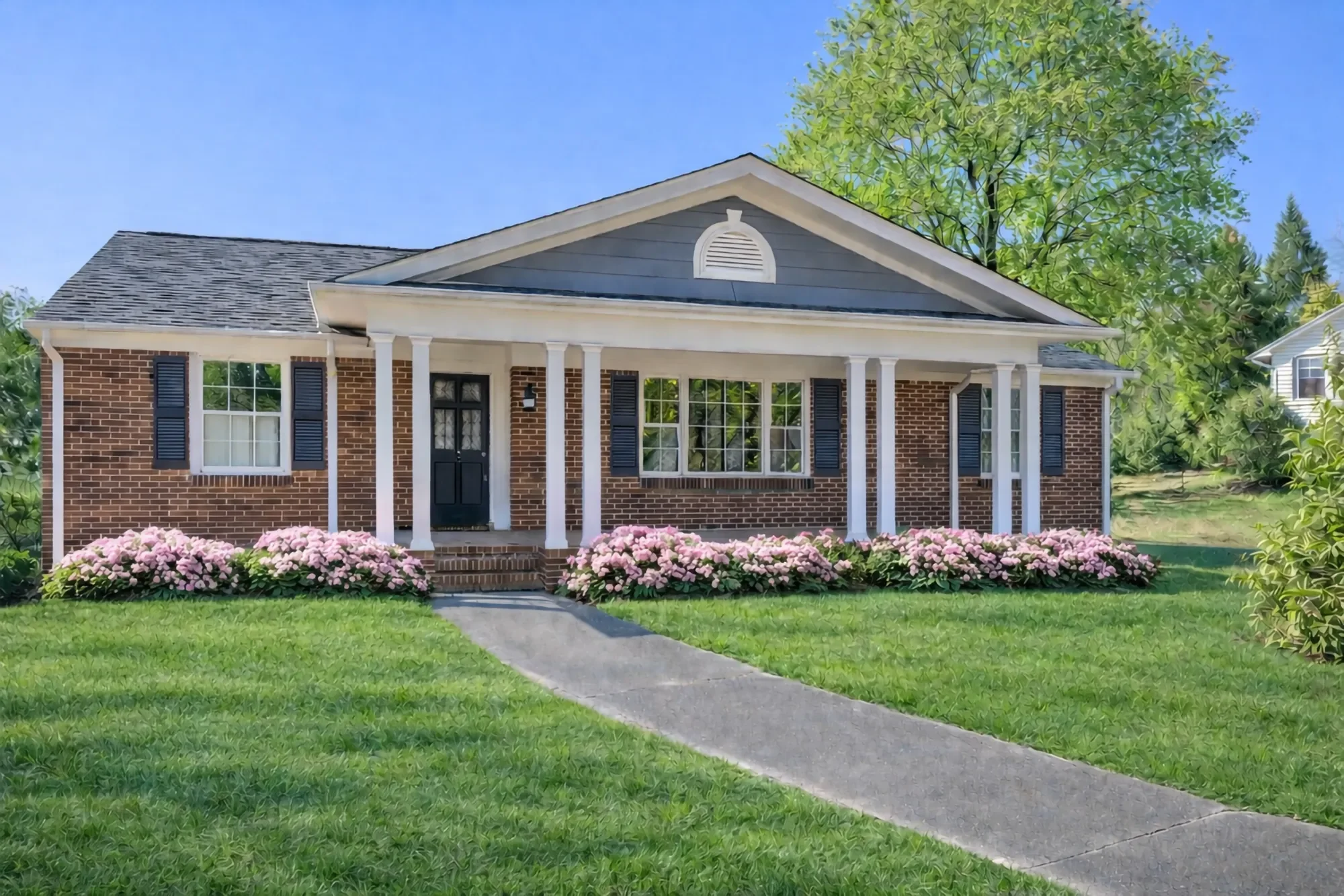 Front view of a brick house with a black door, white columns, and pink flowers in the yard during daytime.