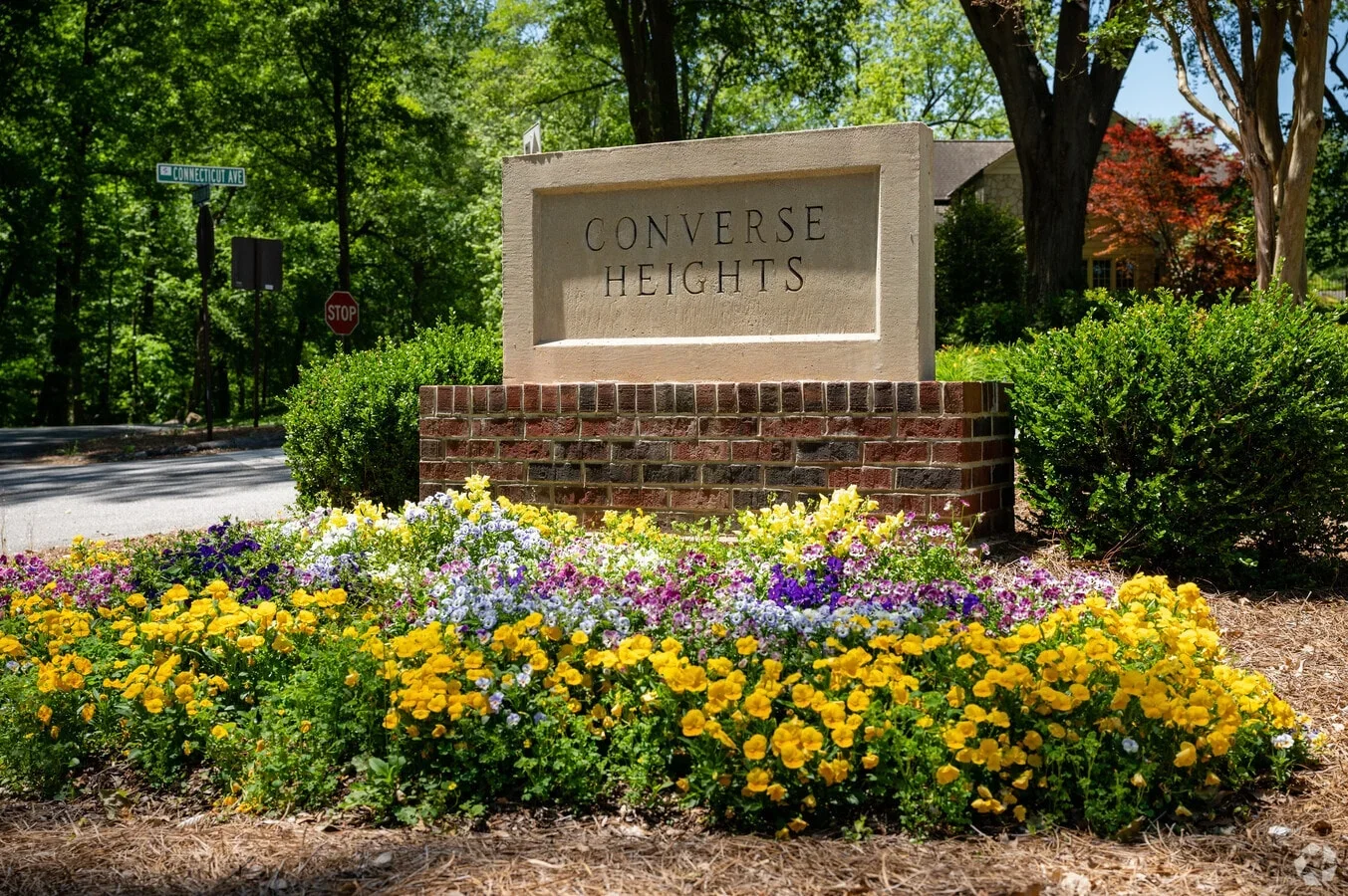 A stone and brick sign reading 'Converse Heights' surrounded by yellow, purple, white, and pink flowers, with trees and a street sign in the background.