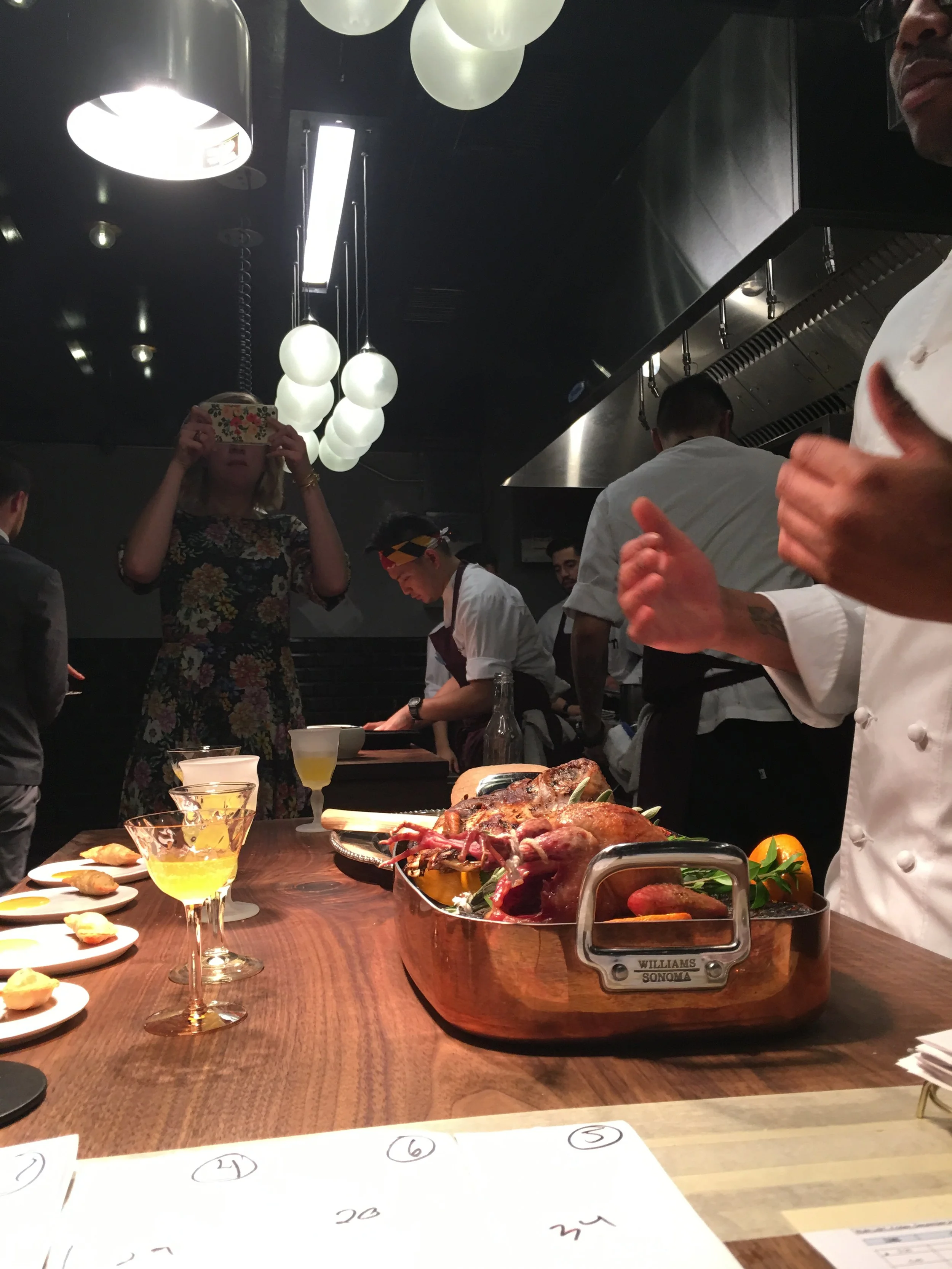 A chef's knife and cooked meat on a wooden cutting board, with appetizers and desserts on small plates, in a restaurant kitchen with chefs and a woman taking a photo.