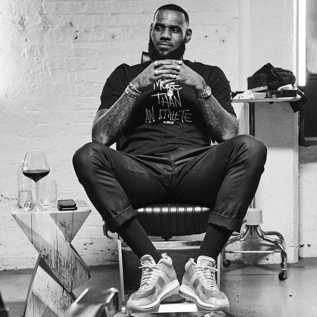 A man with tattoos sitting in a chair with legs crossed, wearing sneakers, a watch, and a T-shirt with text, with a glass of red wine on a small table beside him, in a room with a white brick wall.