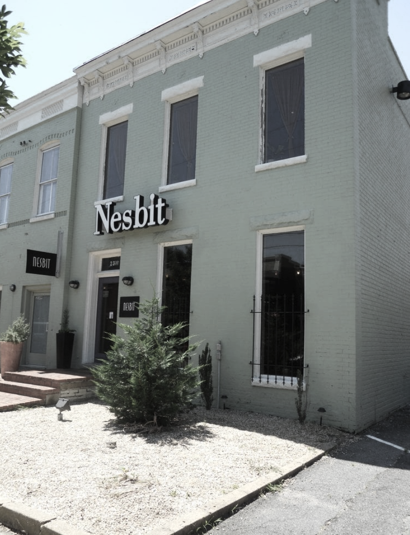 Exterior view of a light green brick building with black and white signage that reads 'Nesbit' at the entrance, with several windows and some small plants or bushes in front.