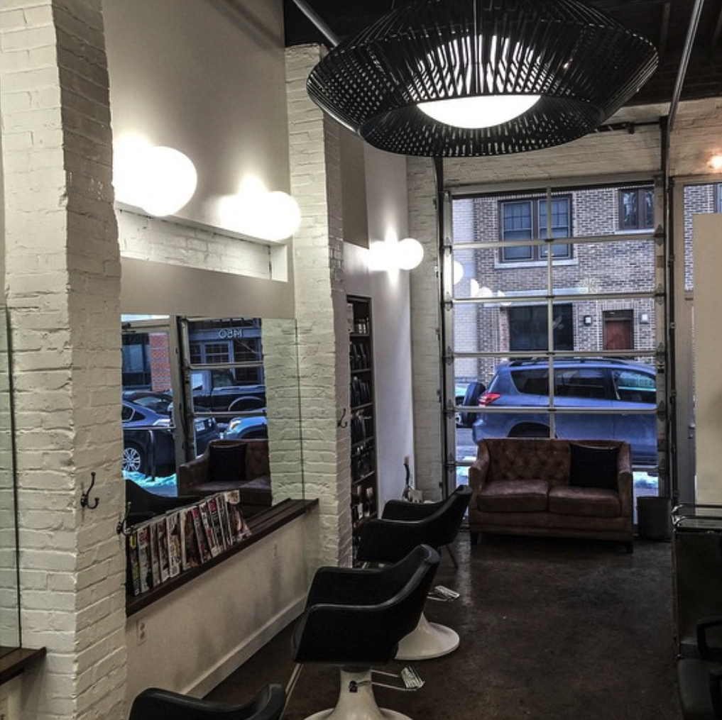 Interior of a modern hair salon with salon chairs, a large window, and a leather couch, featuring brick walls and decorative lighting.