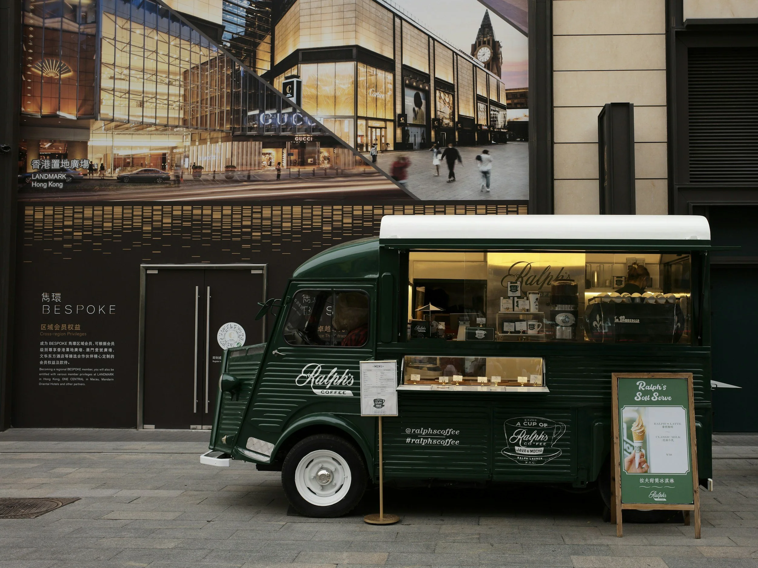 A green vintage-style mobile coffee cart with a white roof, displaying menu signs and branding for Ralph's Coffee, set on a city sidewalk with an billboard above advertising luxury stores like Gucci in Hong Kong.