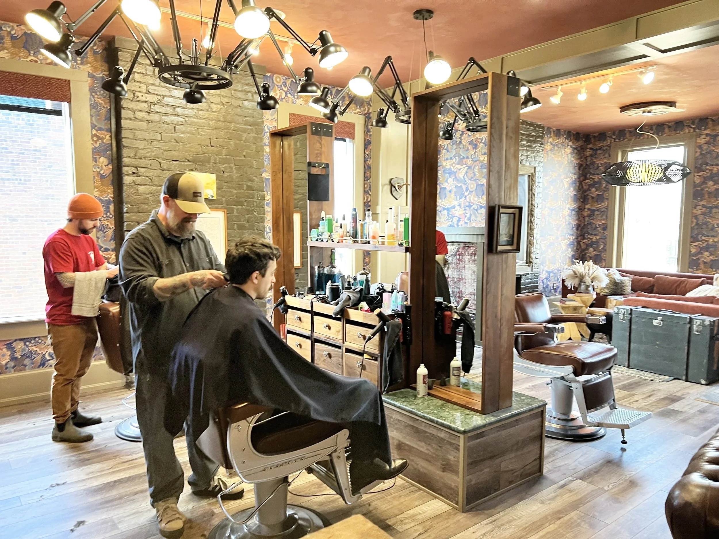 Barber shop with a barber cutting a young man's hair and another barber preparing to cut hair in the background, inside a cozy lounge with vintage wallpaper, wooden furniture, and large windows.