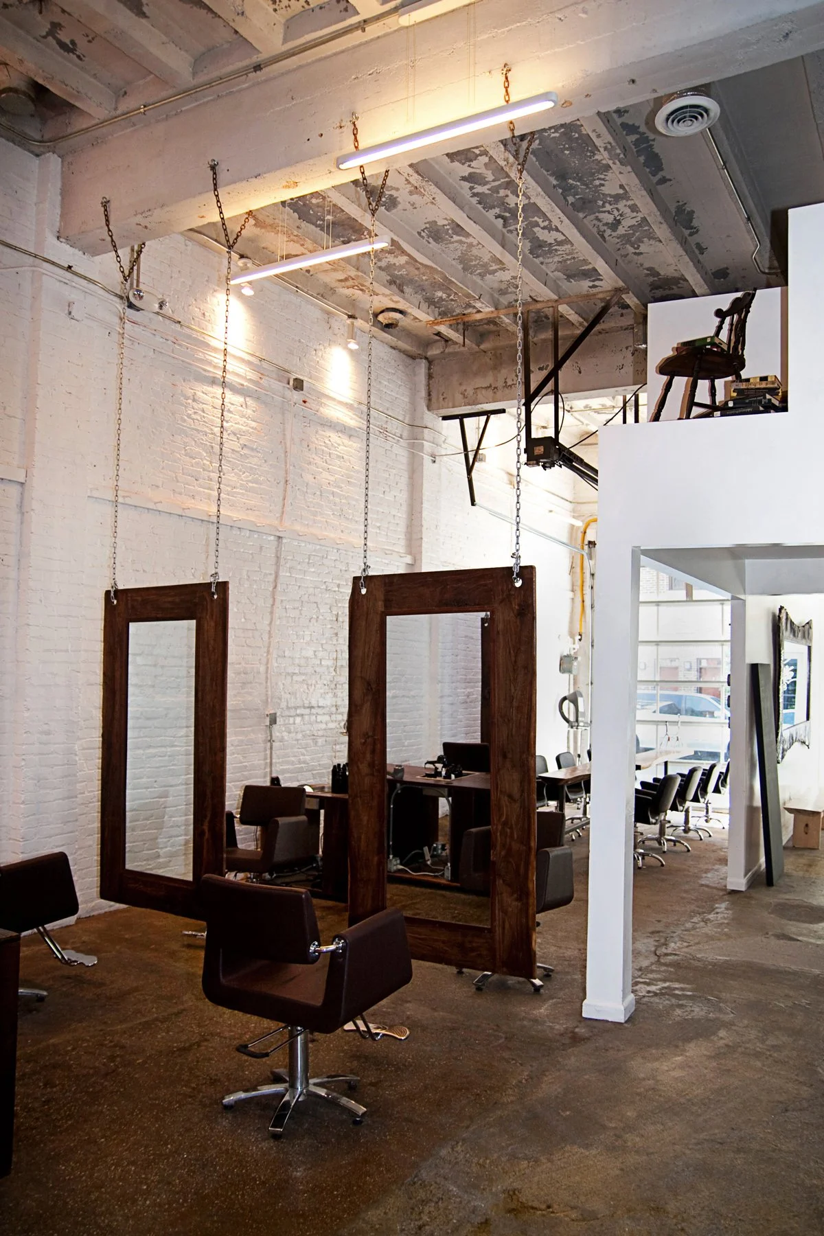 An industrial-style hair salon with exposed ceiling beams, hanging light fixtures, and large mirrors in wooden frames. Hair styling chairs are arranged in the space, and a small upstairs nook with books and a chair is visible.