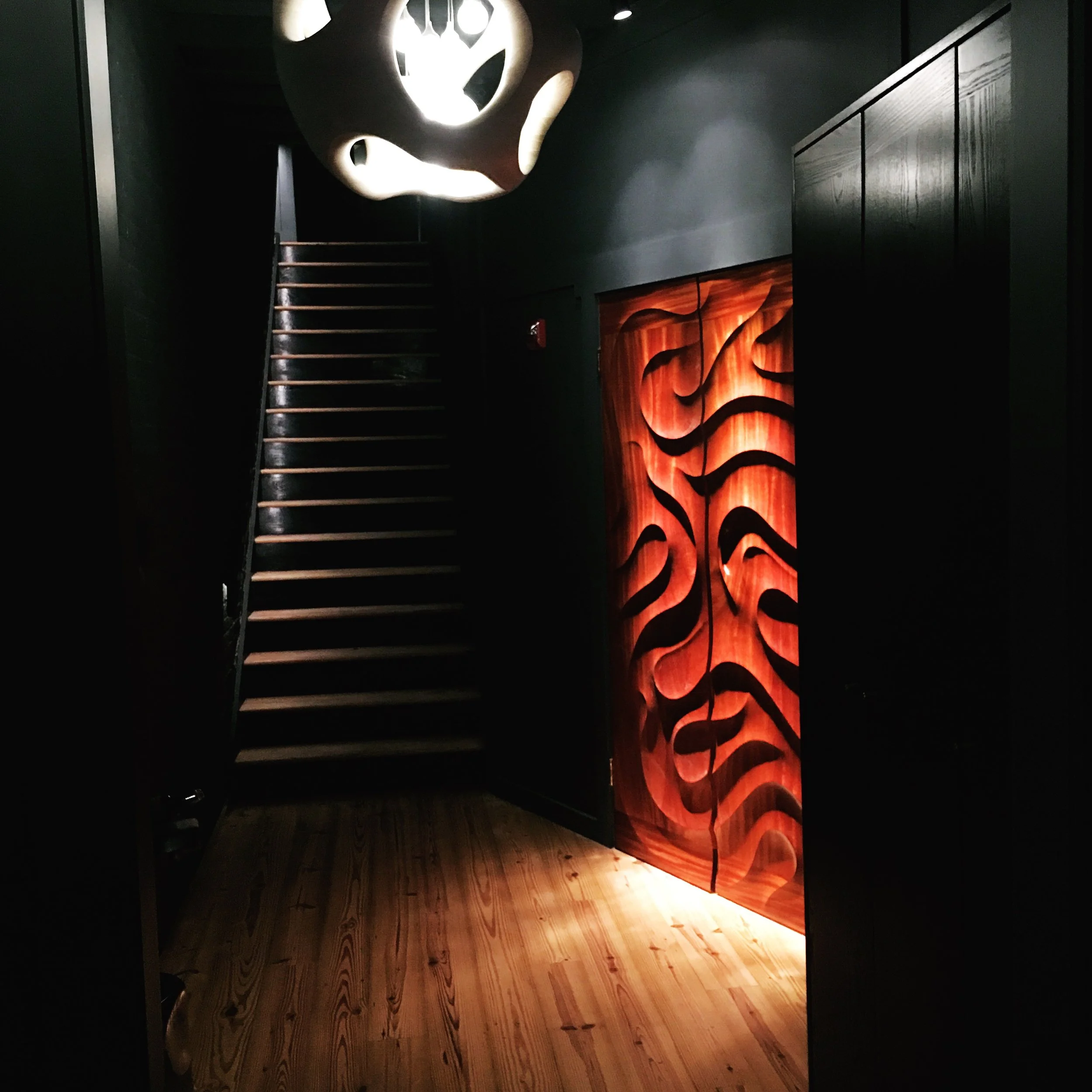 Dark interior staircase with a wooden railing, illuminated artwork with fiery red and orange swirling patterns on a wall or door, and an abstract light fixture overhead.