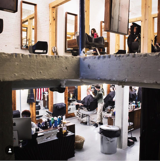 View of a busy barbershop with multiple patrons getting haircuts, barbers working, and mirrors on the walls.