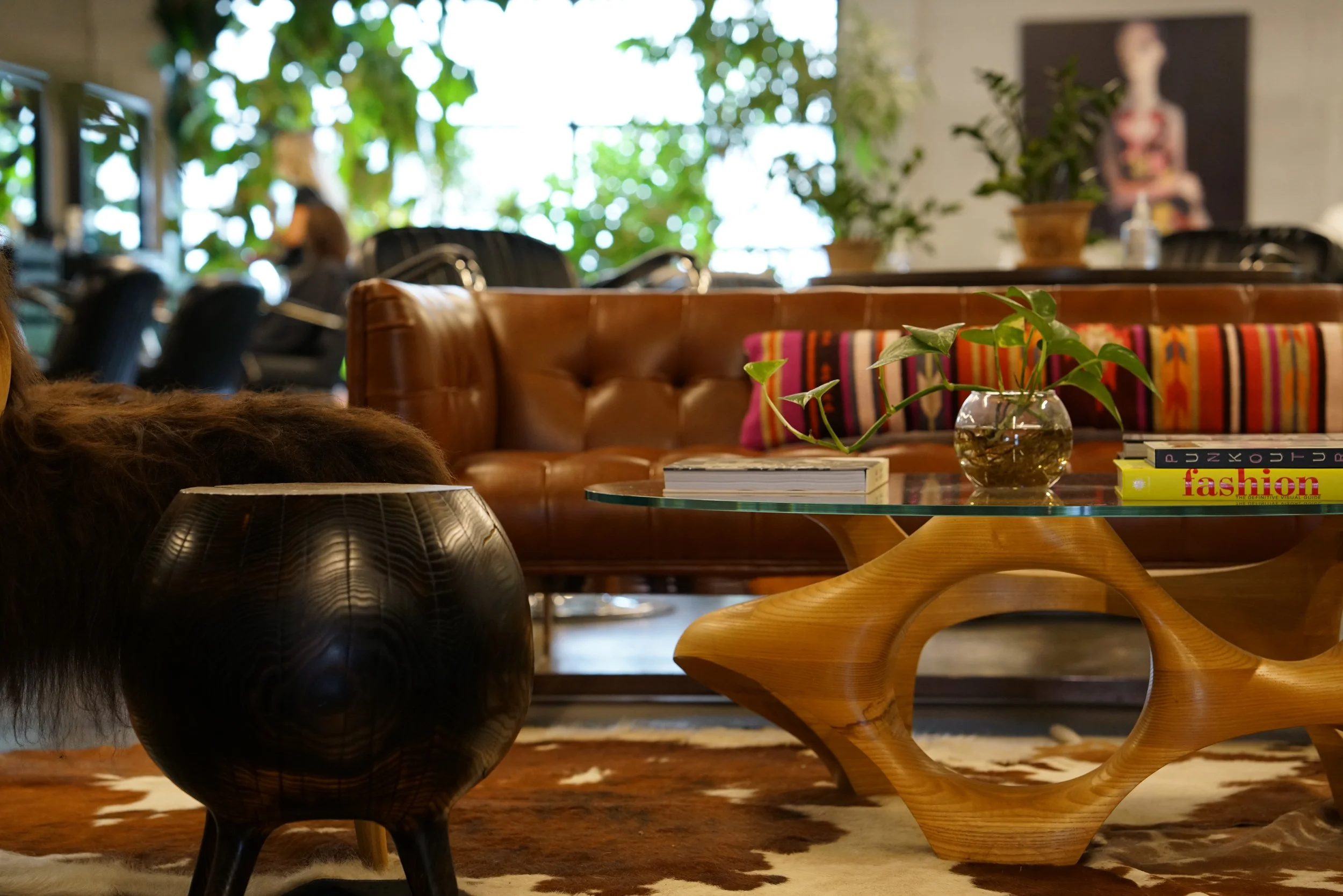 A stylish modern living room with a glass-top coffee table, a brown leather sofa with colorful striped pillows, a glass vase with green plants, and a stack of books, including one titled 'fashion.' In the background, there are blurred figures and lus