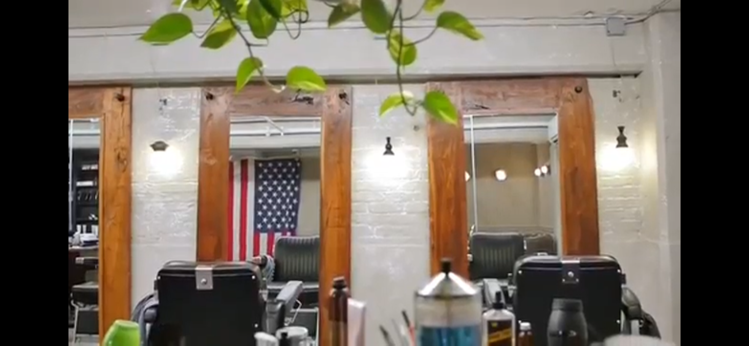 Interior of a barber shop or salon with barber chairs facing a mirror wall, decorated with wooden panels and an American flag hanging in the background.
