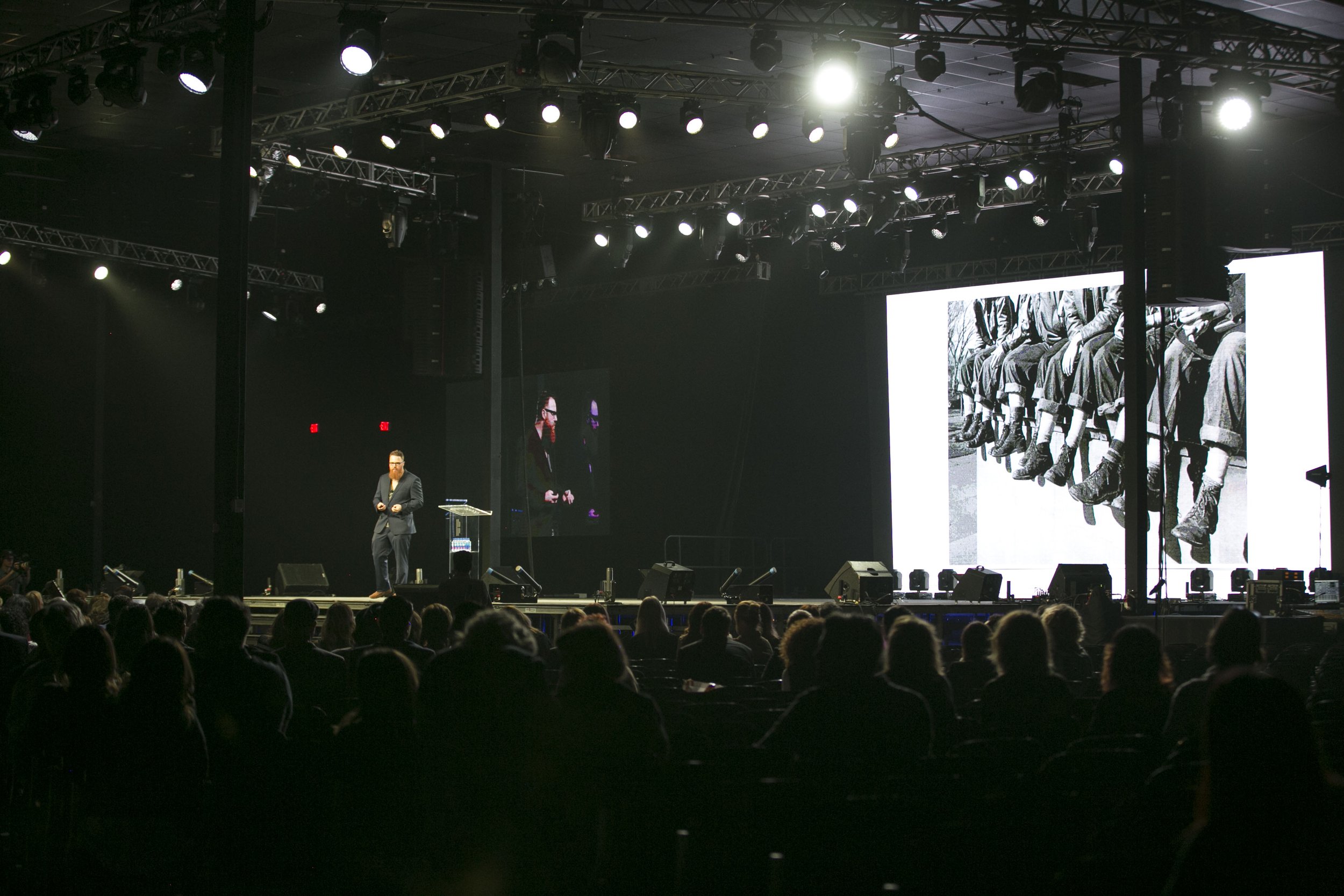 A speaker presenting on a stage at a conference or event, with large screens behind him showing black-and-white historical photo of men sitting on a ledge. The audience is seated in front of the stage in a darkened room.
