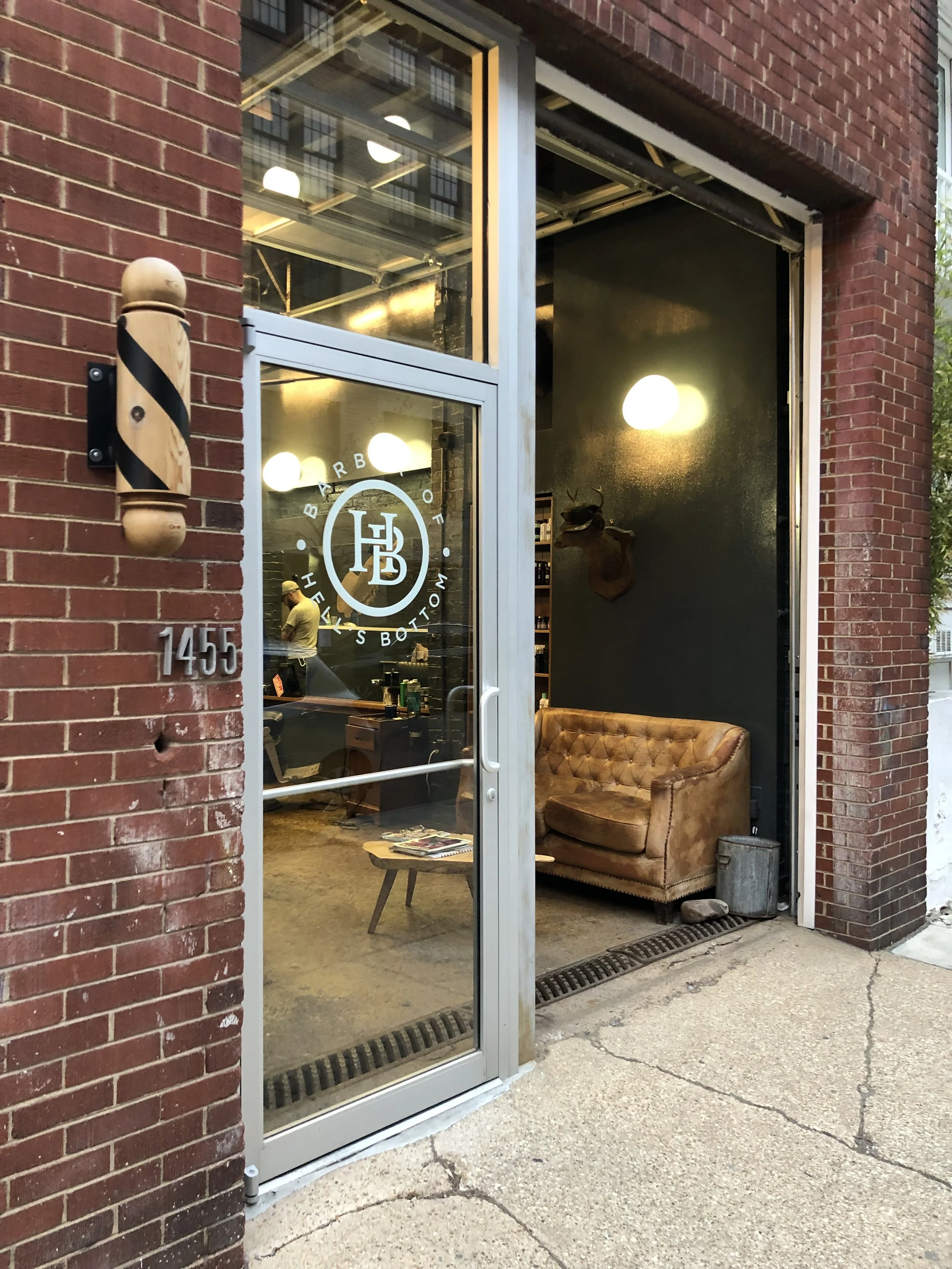 The exterior of a barber shop with a glass door displaying the logo 'Barbershop of WLM' and the street number 1455. Inside, a cozy waiting area has a vintage leather sofa and a mounted animal head on the dark wall, with warm lighting overhead.