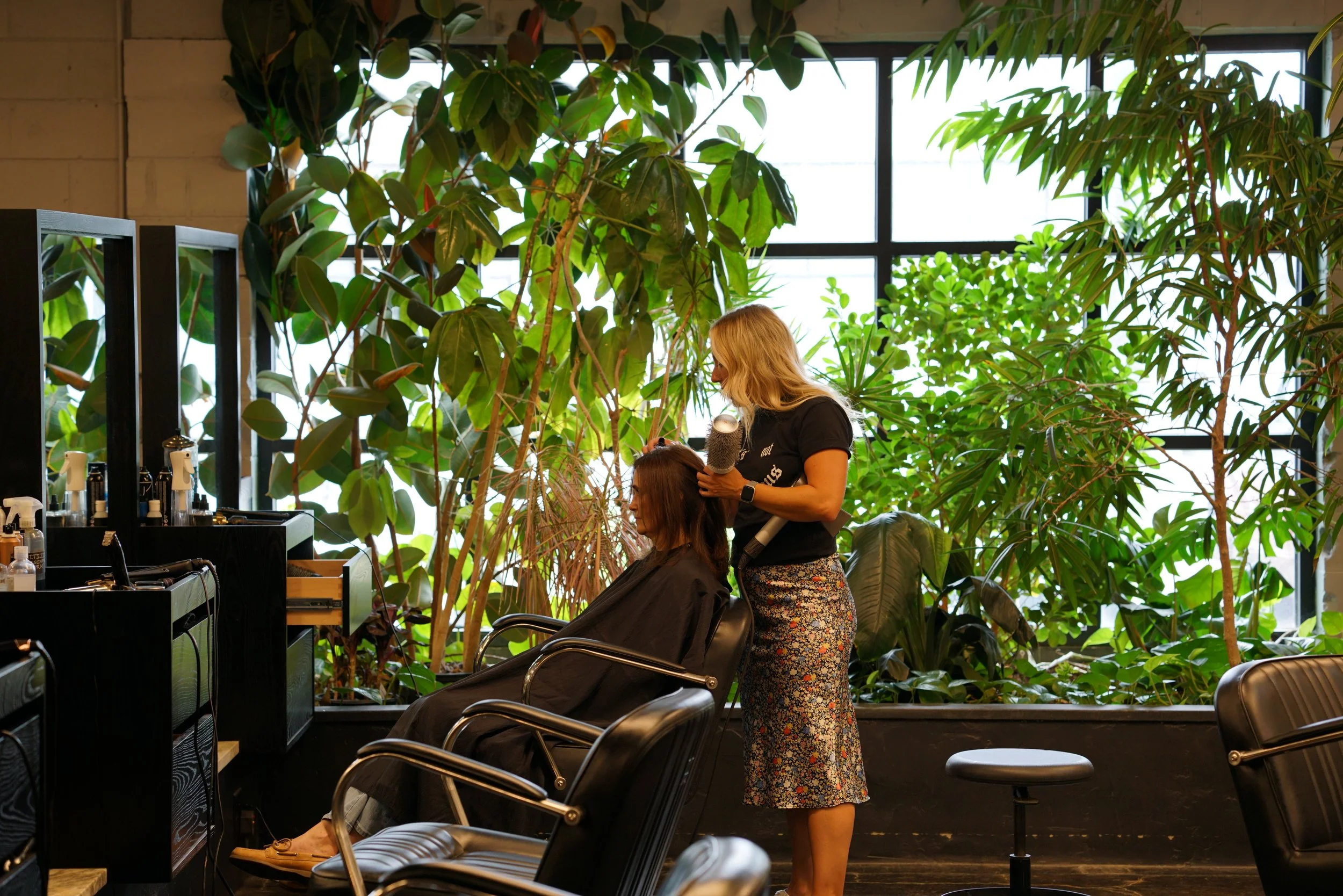 A woman is getting her hair styled by a hairstylist in a salon with large green plants and big windows in the background.