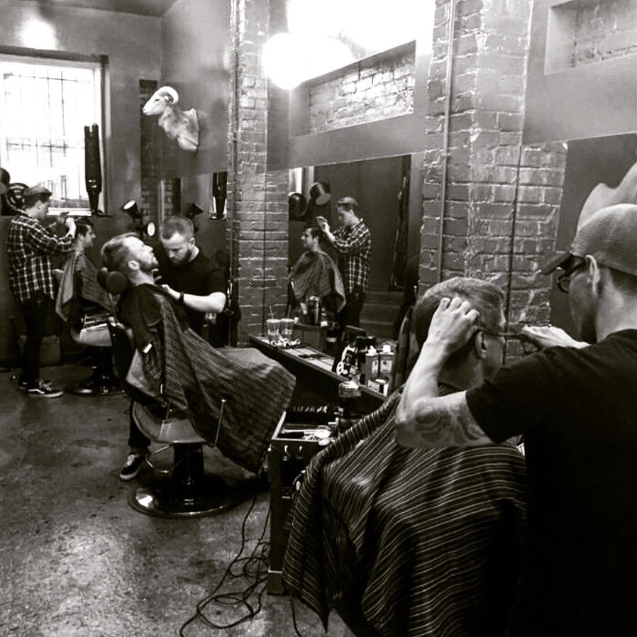 Barbershop interior with multiple barbers cutting customers' hair, brick walls, and a mounted animal head on the wall.