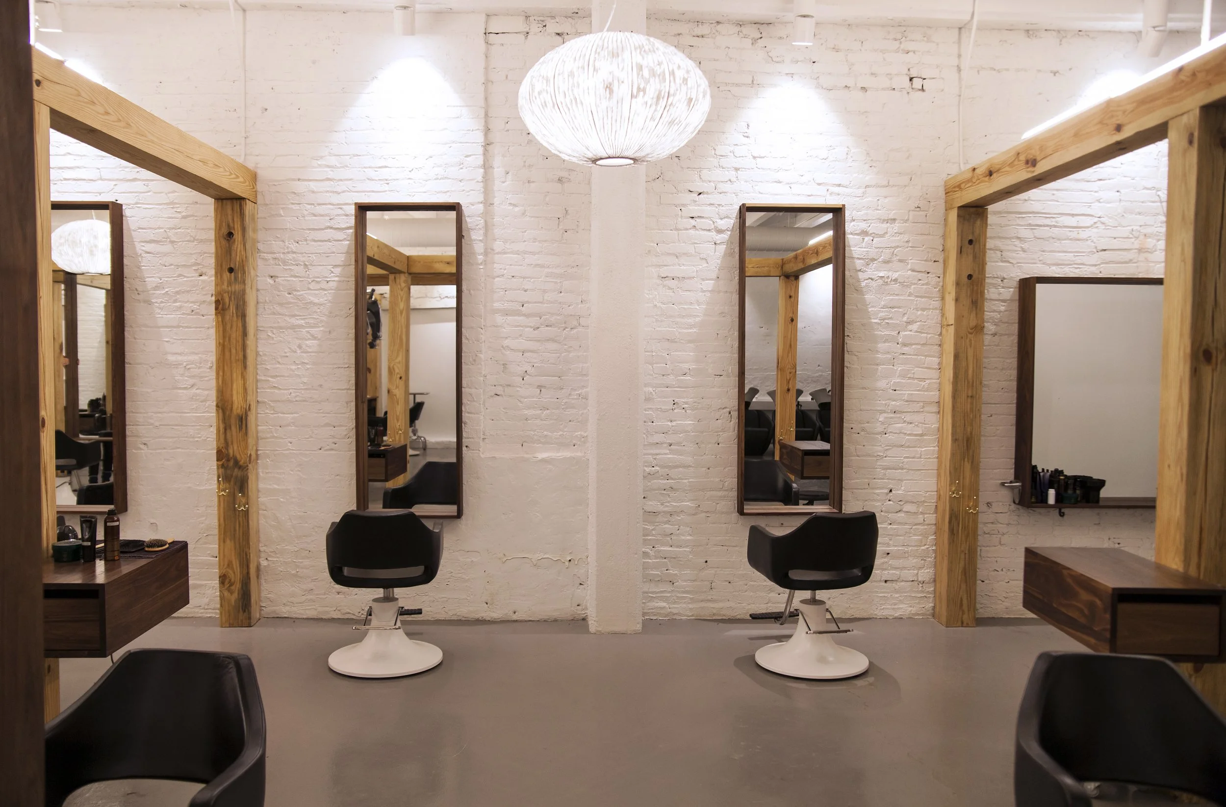 Empty hair salon with two styling stations, each with a mirror, black chair, and wooden frame, set against a white brick wall with a central pillar, ceiling lights, and a pendant lamp.