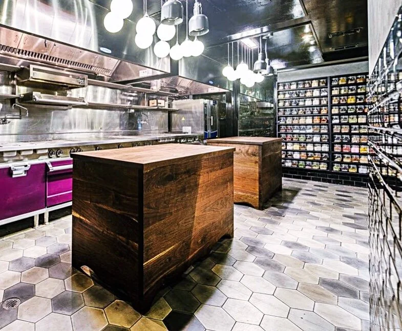 Empty modern kitchen with wooden island, hexagonal tile flooring, and wall of shelves filled with jars or containers.