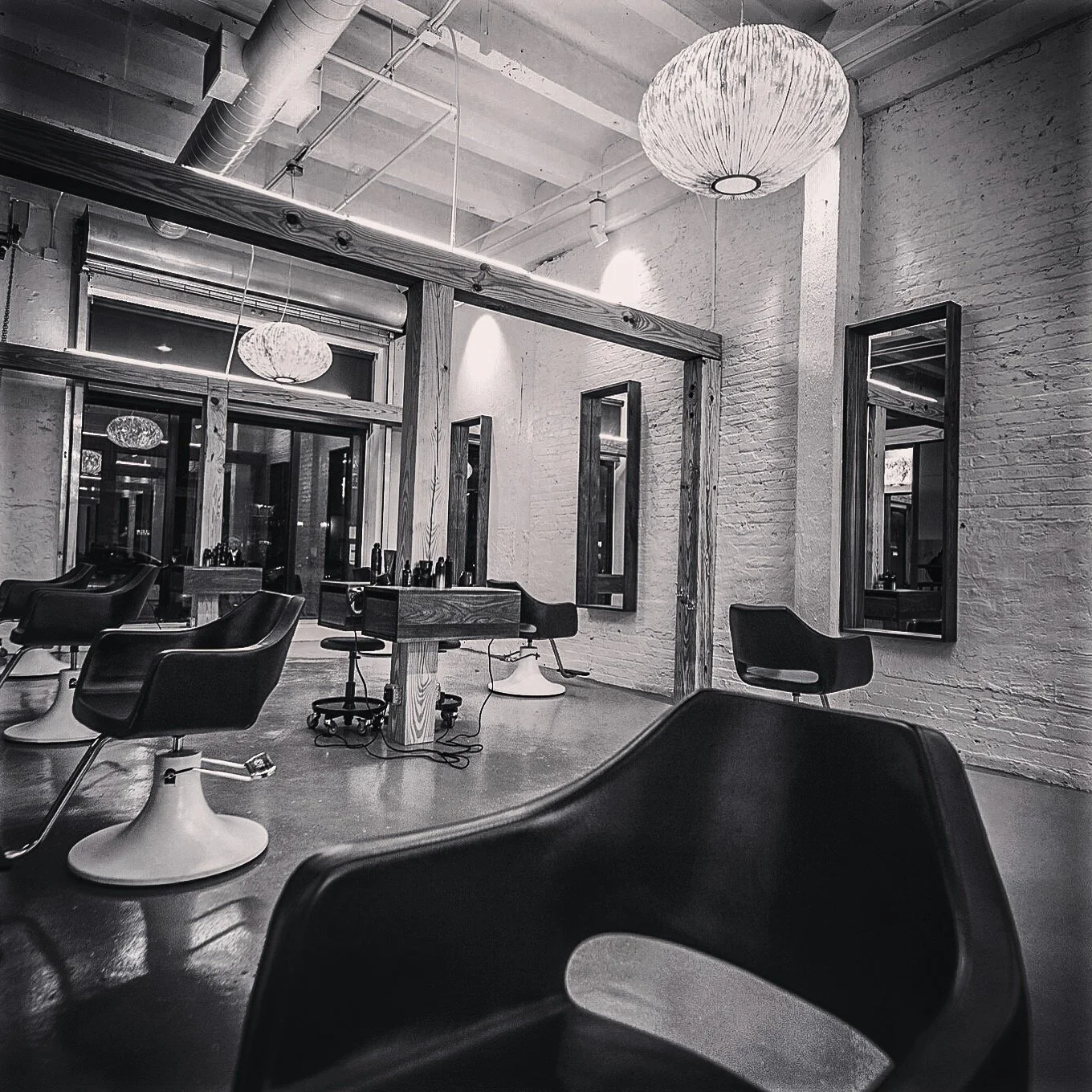 Interior of a modern hair salon with black salon chairs, mirrors on brick walls, and hanging ceiling lights in black and white.