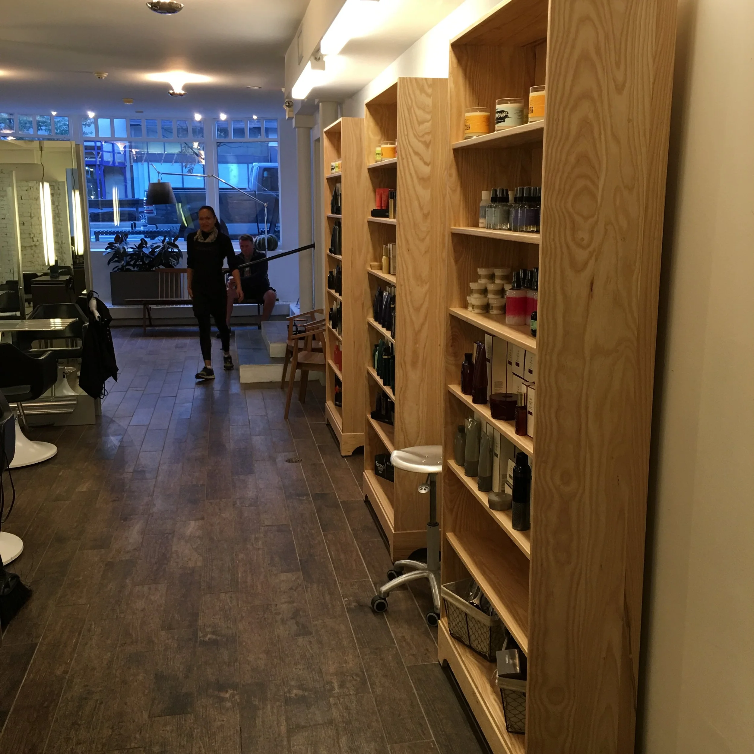 Interior of a store or salon with wooden shelves displaying various beauty or hair products, a stool, and people walking and sitting in the background.