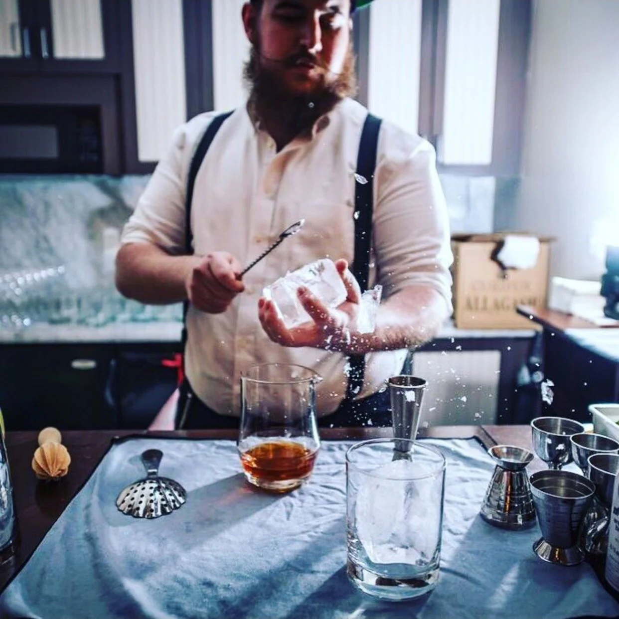A man with a beard wearing a white shirt and suspenders is breaking an ice cube with a bar spoon over a bar counter. There are various cocktail-making tools, glassware, and a drink in a glass on the counter.