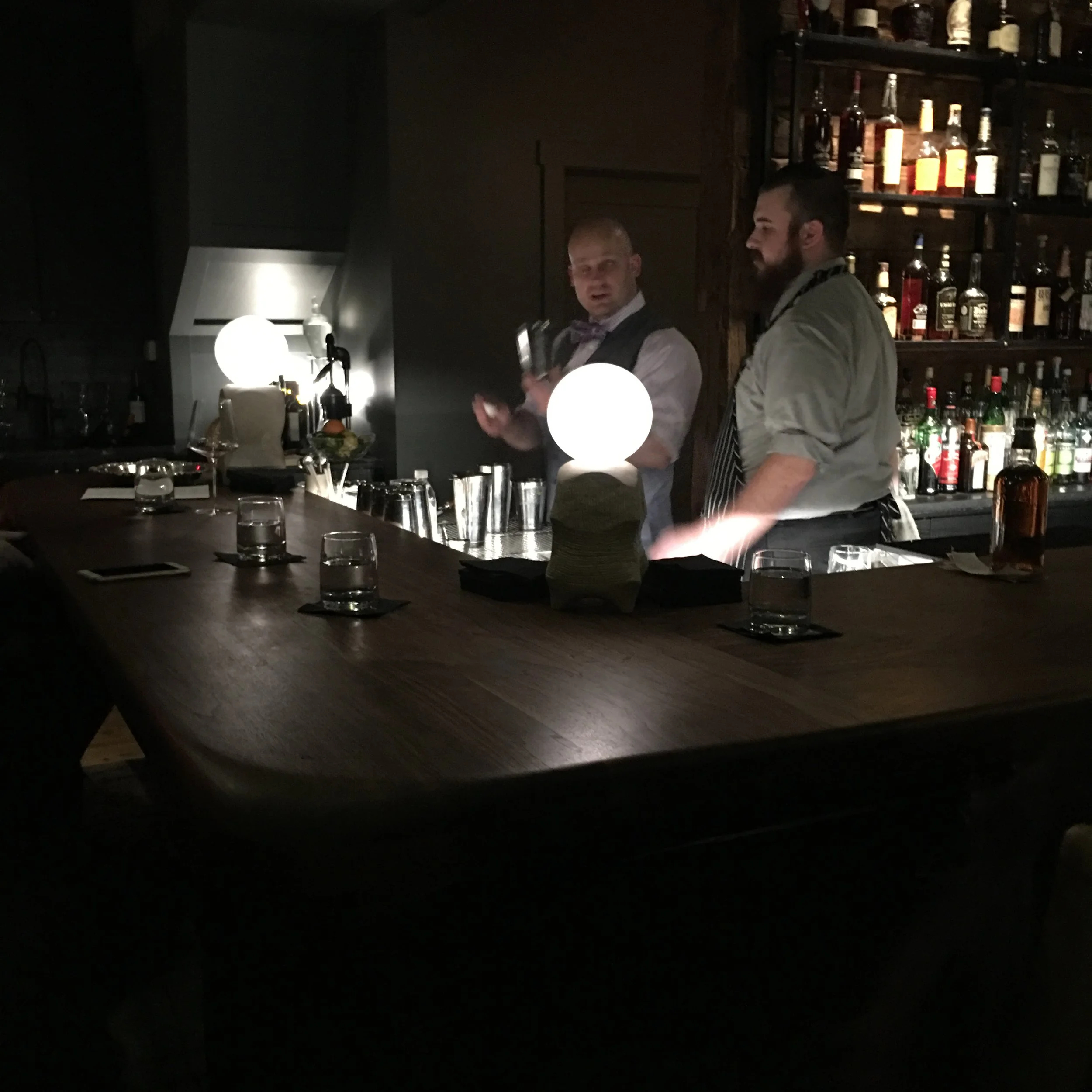 Two bartenders behind a bar counter in a dimly lit bar or restaurant, with shelves of liquor bottles in the background. One bartender is holding a cocktail shaker, and the other is standing nearby, engaged in conversation or preparing drinks.
