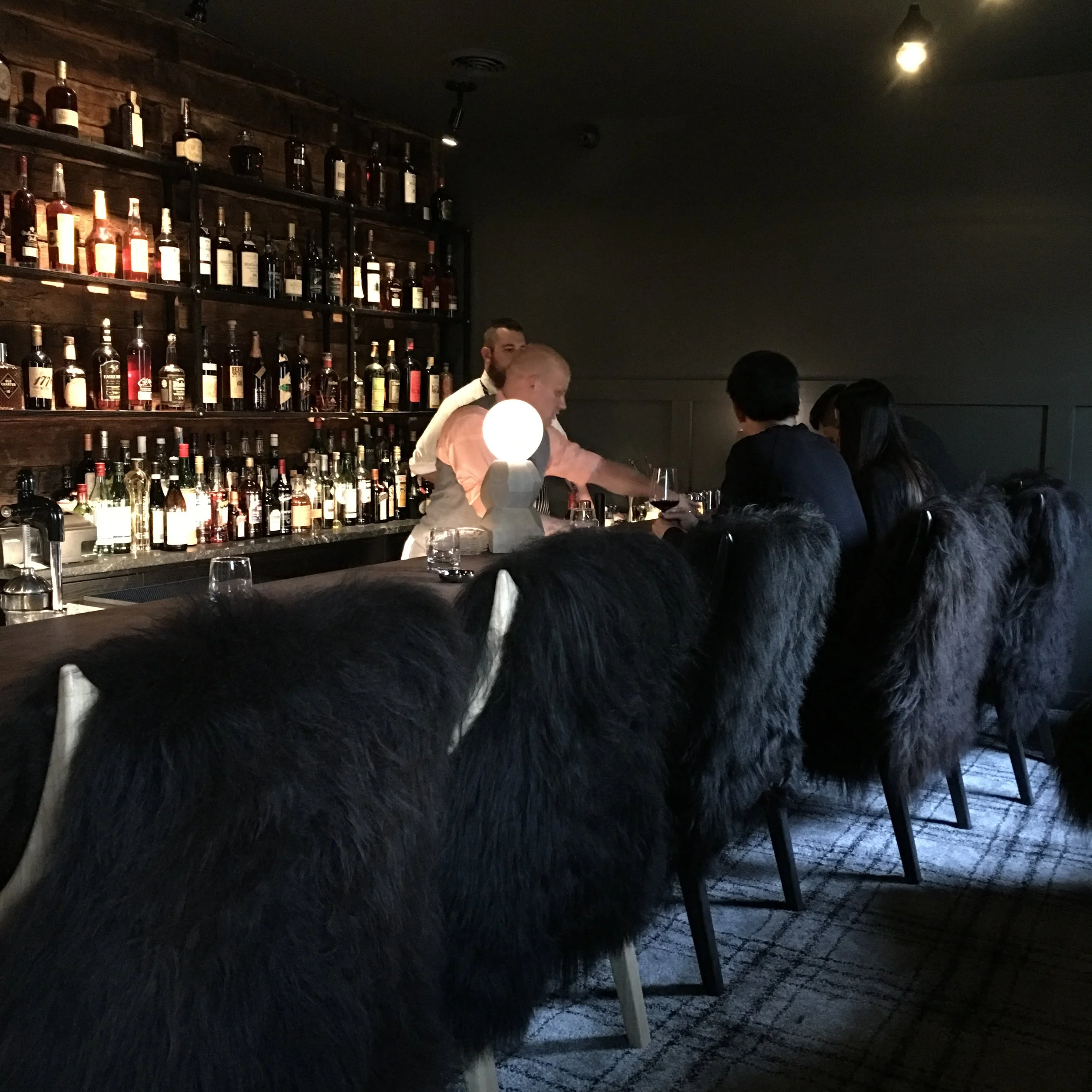 Bar with people sitting at the counter, bartender serving drinks, shelves filled with liquor bottles, and chairs with black fur covers.