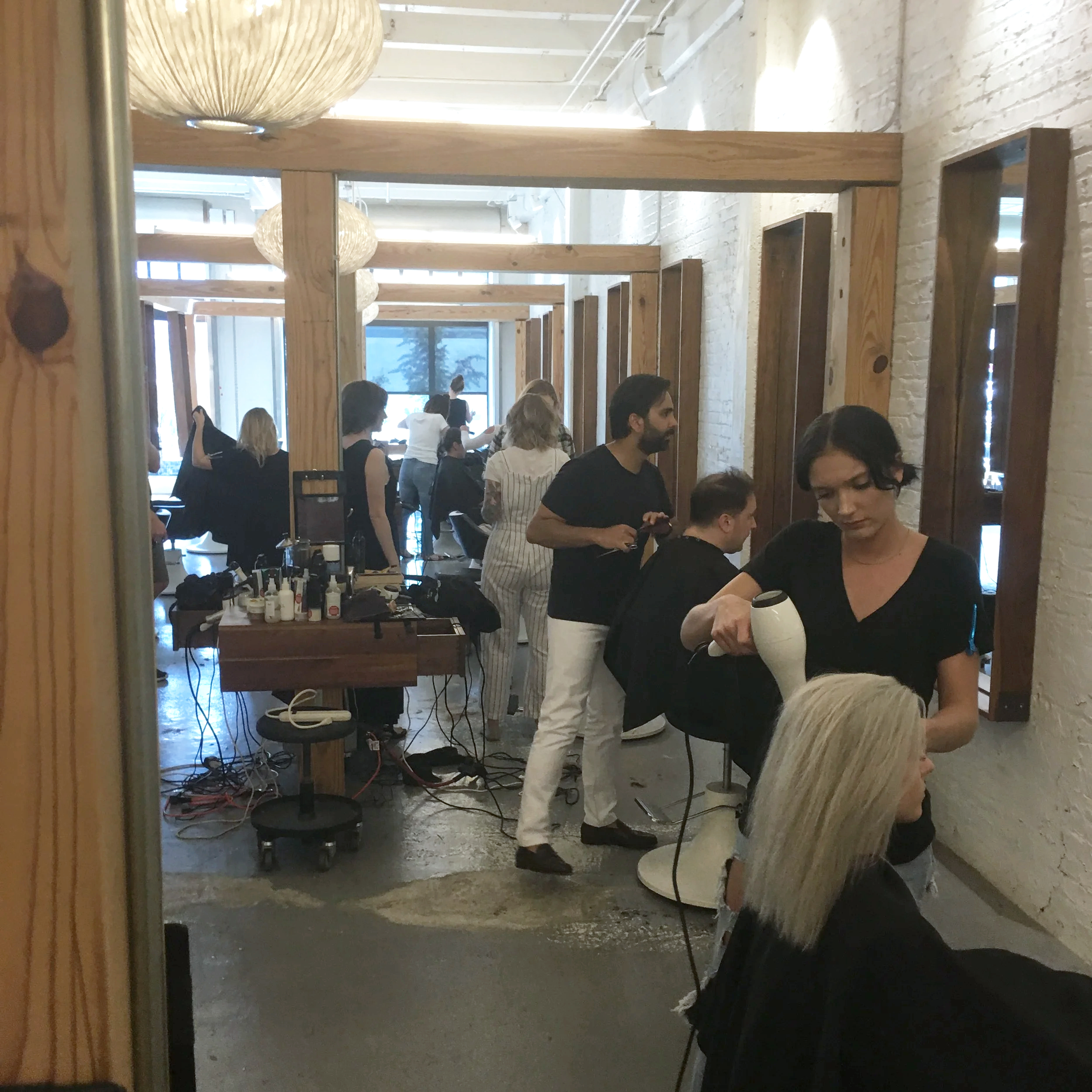Hair stylists working in a busy salon, with clients in chairs and hairdressing tools on a table, and multiple people standing and working in the background.