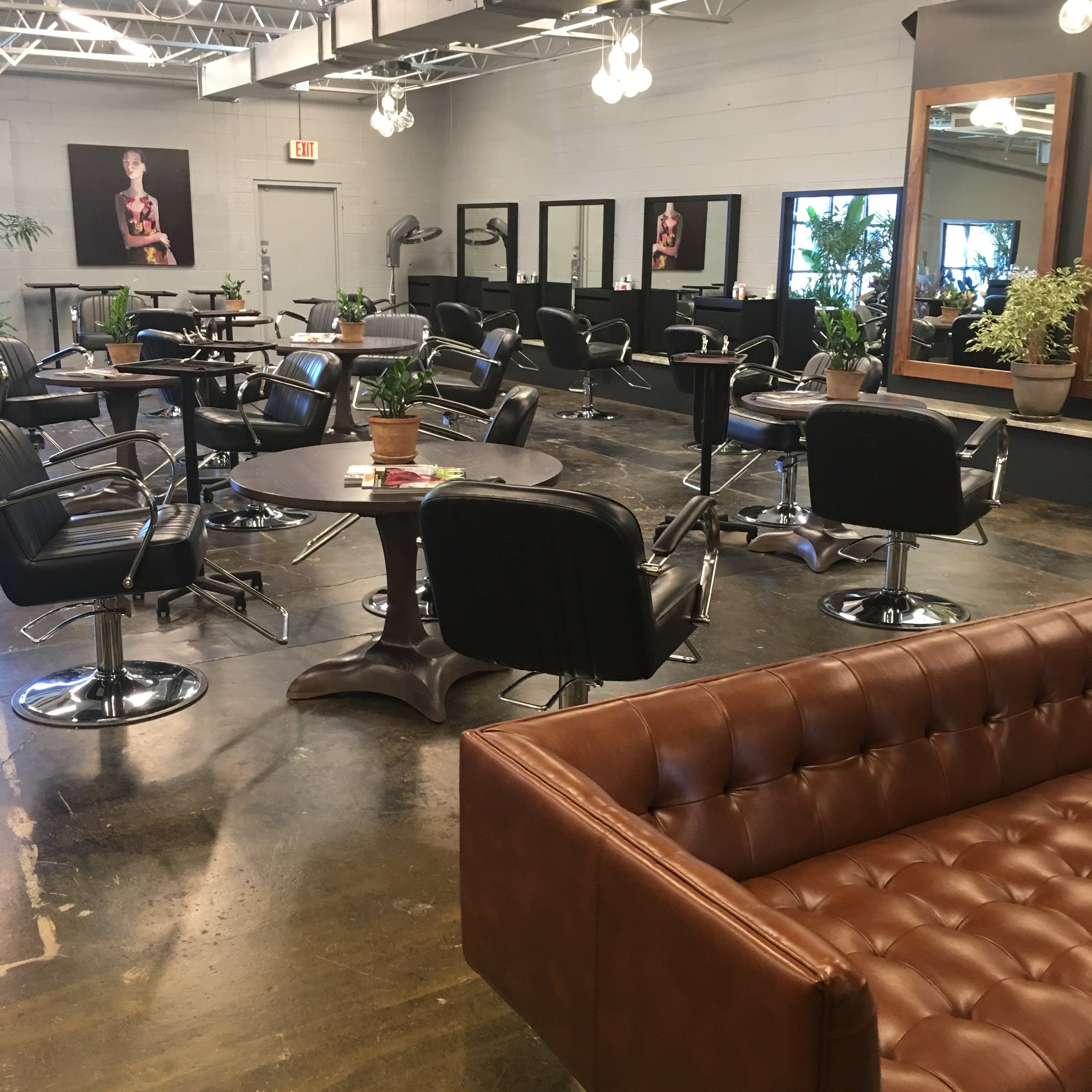 A stylish hair salon with black salon chairs, round tables with potted plants, and mirrors along the wall. There is a brown leather tufted sofa in the foreground, and a gray wall with artwork. The salon has modern lighting and a polished concrete floor.
