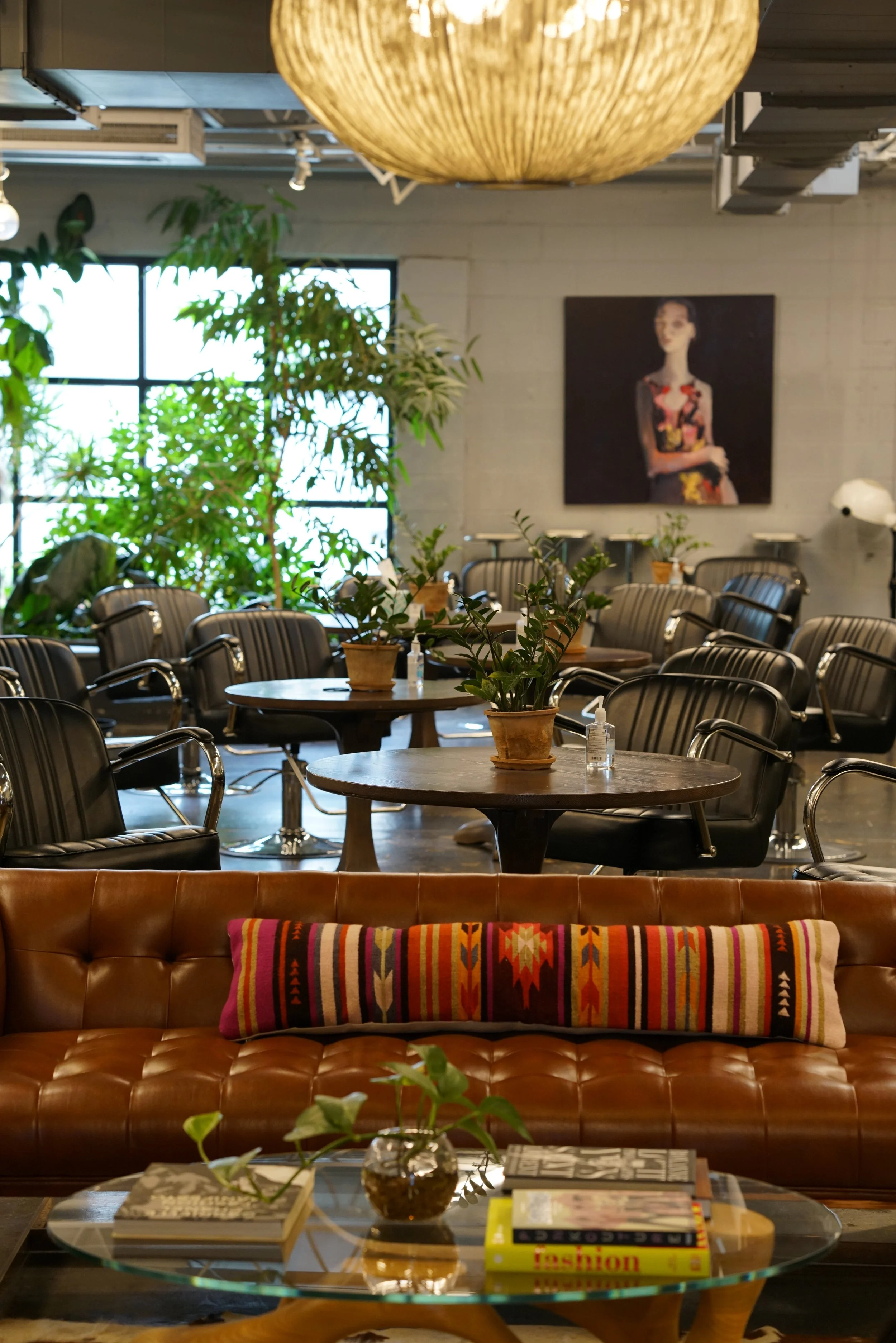 Cozy, stylish coffee shop with a leather couch, colorful pillow, glass coffee table with books and a plant, wooden tables with plants, black chairs, large window, indoor plants, wall art, and ambient lighting.