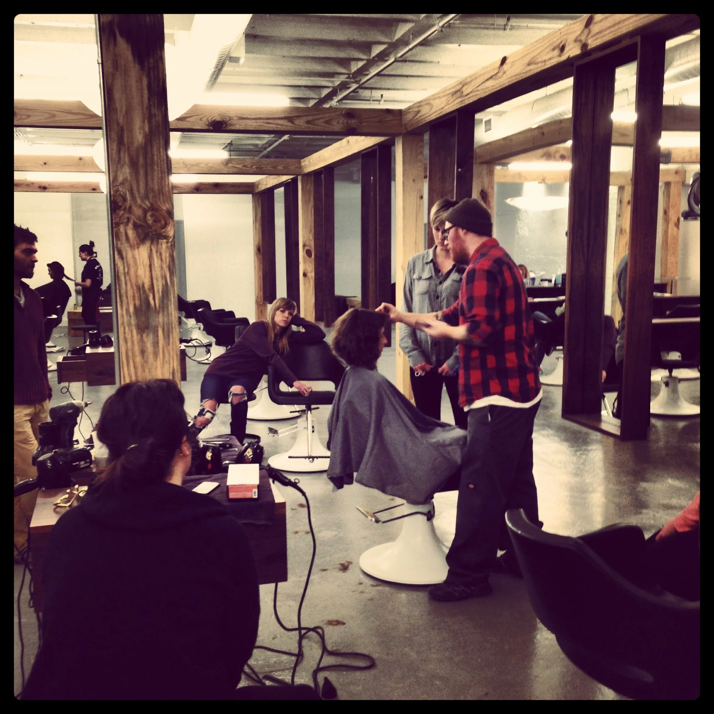 A hairstylist in a red plaid shirt demonstrates a haircut to a woman seated in a salon chair, while two people observe nearby; others are waiting or working in the salon, which has a modern industrial design with wooden beams.