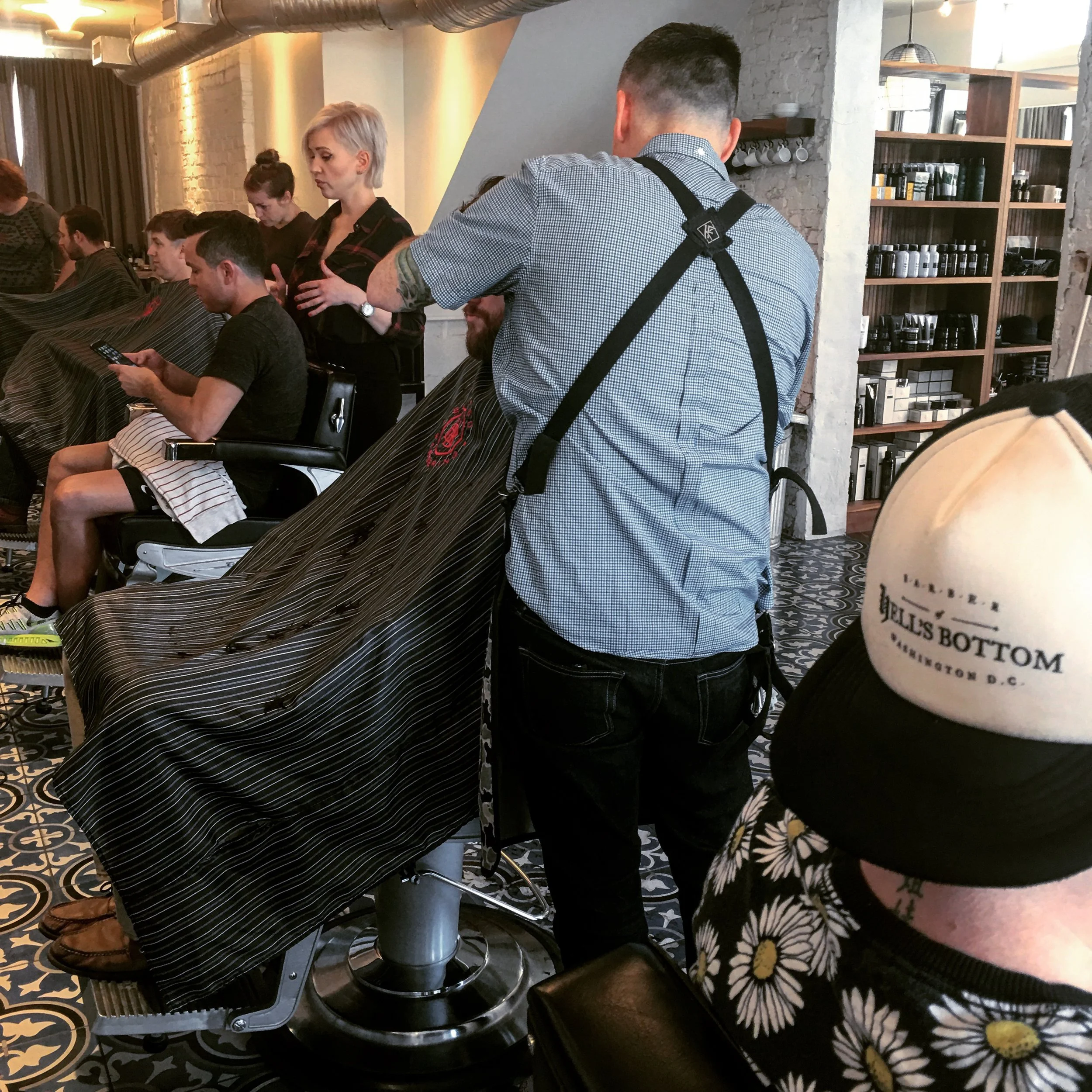 A barber in a checkered shirt gives a haircut to a man seated in a barber chair inside a modern barbershop, with customers getting haircuts and a staff member standing in the background.