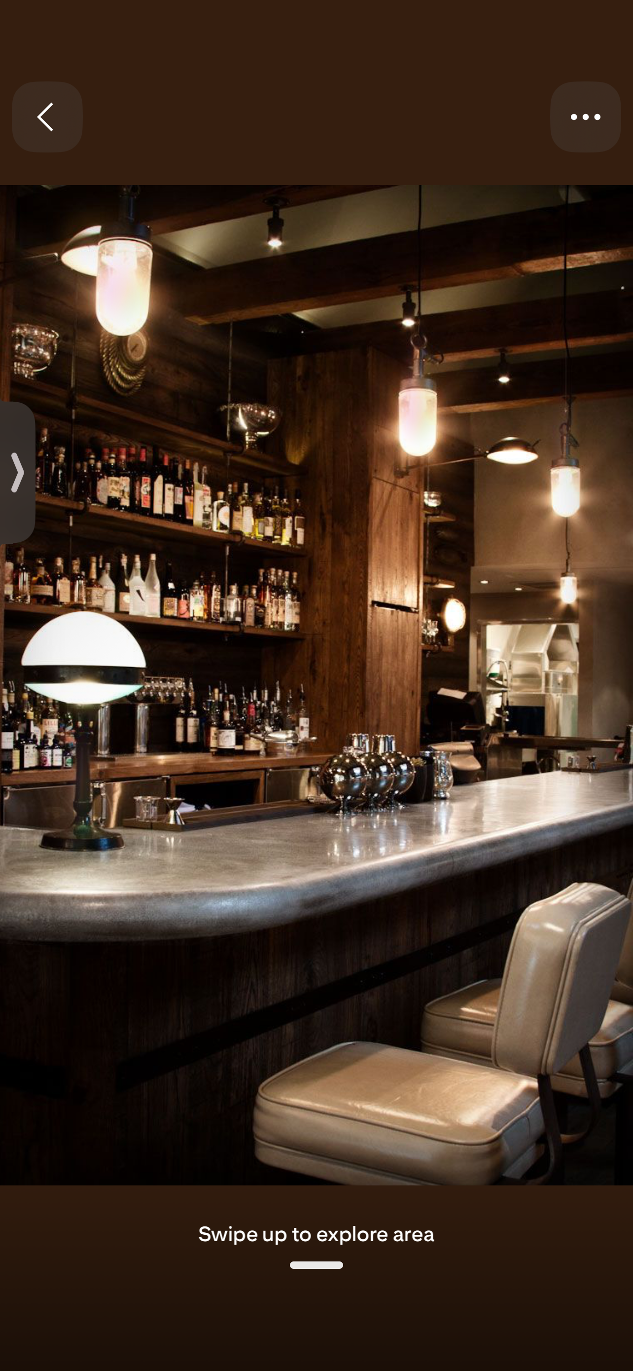 A dimly lit bar with a polished countertop, top shelves filled with various liquor bottles, modern hanging lights, and seating with cream-colored cushions.