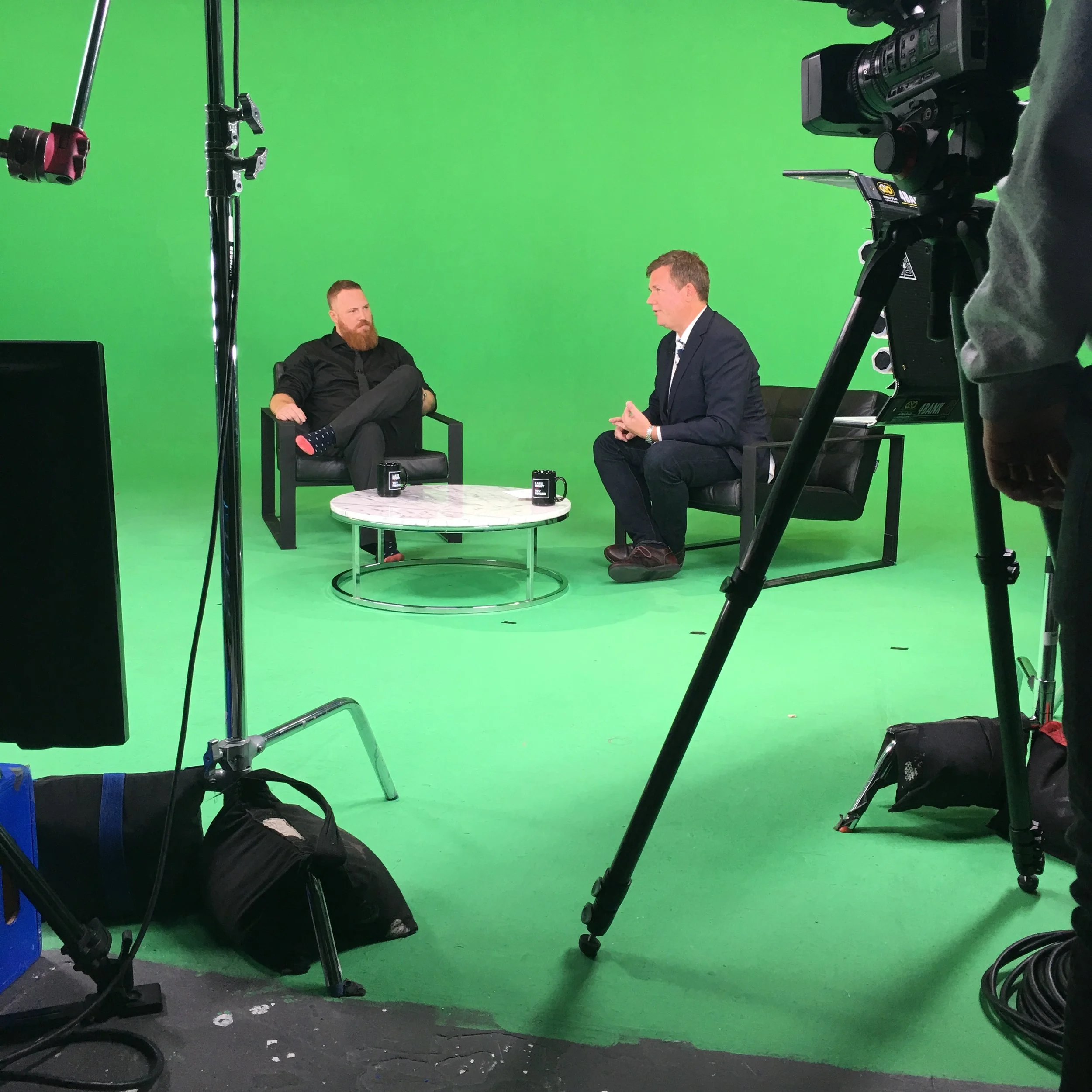 Two men sitting and talking on a TV set with a green screen backdrop, surrounded by filming equipment including cameras, lights, and cables.