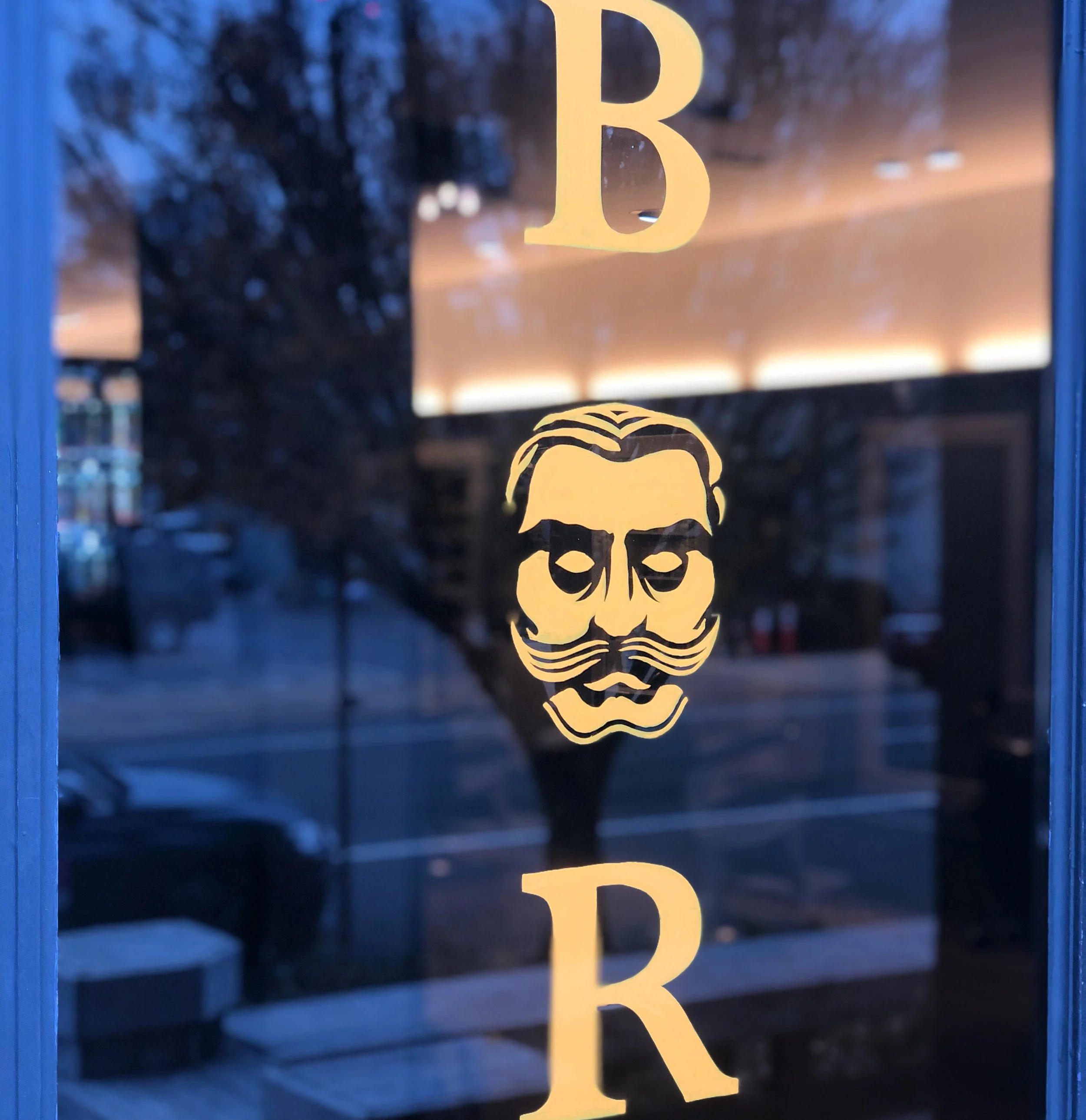 Glass window with a logo of a bearded man with a mustache and hair combed back, with the word 'BAR' vertically arranged around the logo.