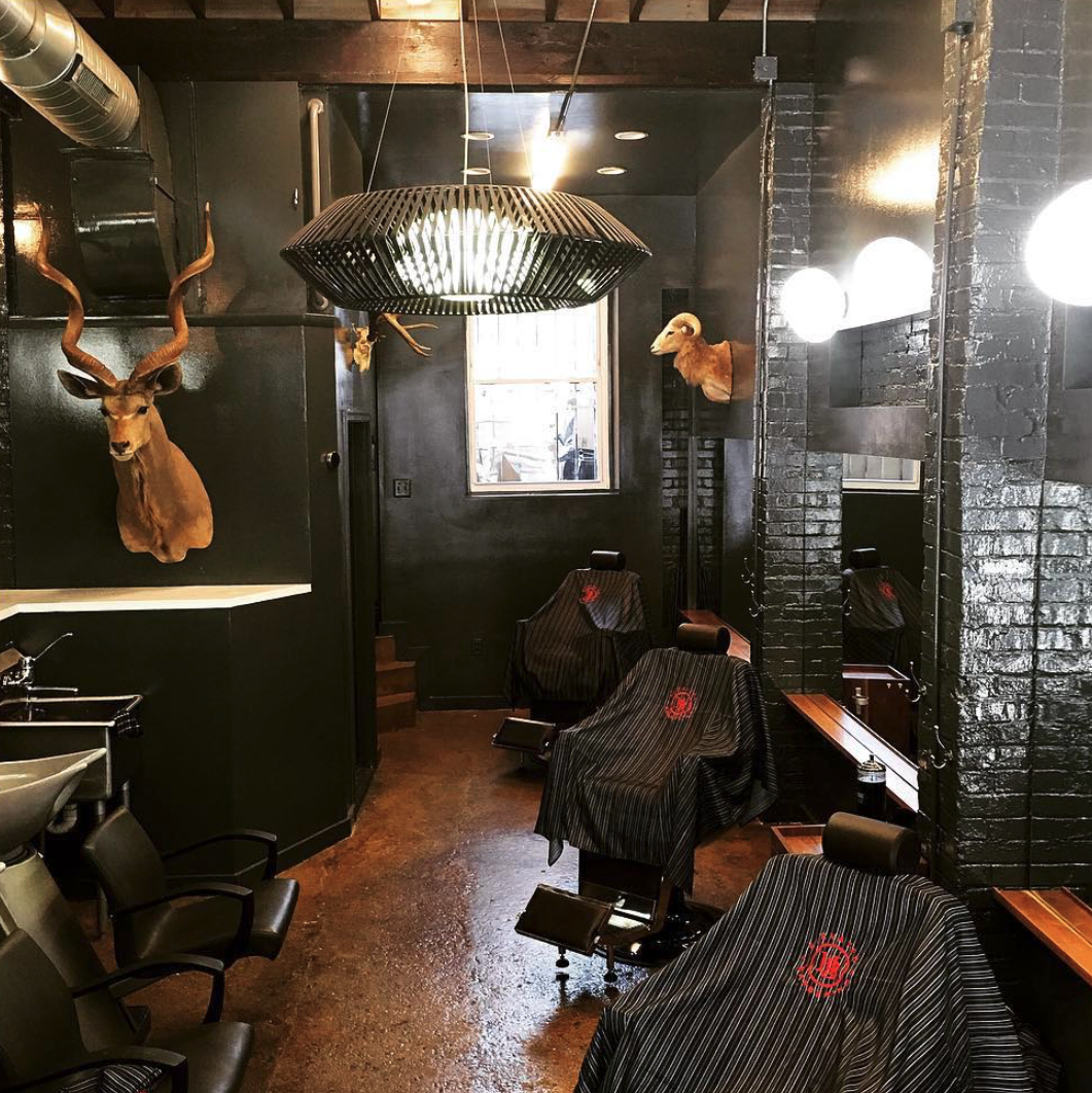 Interior of a barbershop with black painted brick walls, a window, mounted animal heads including a deer and a ram, barber chairs covered with striped cloths, and a large circular pendant light.