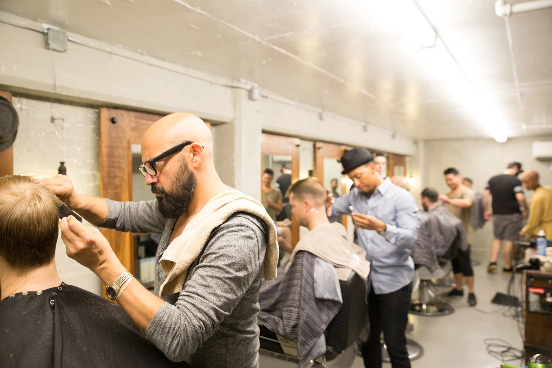 Barbers in a busy salon giving haircuts and styling clients, with people in the background waiting or getting services.