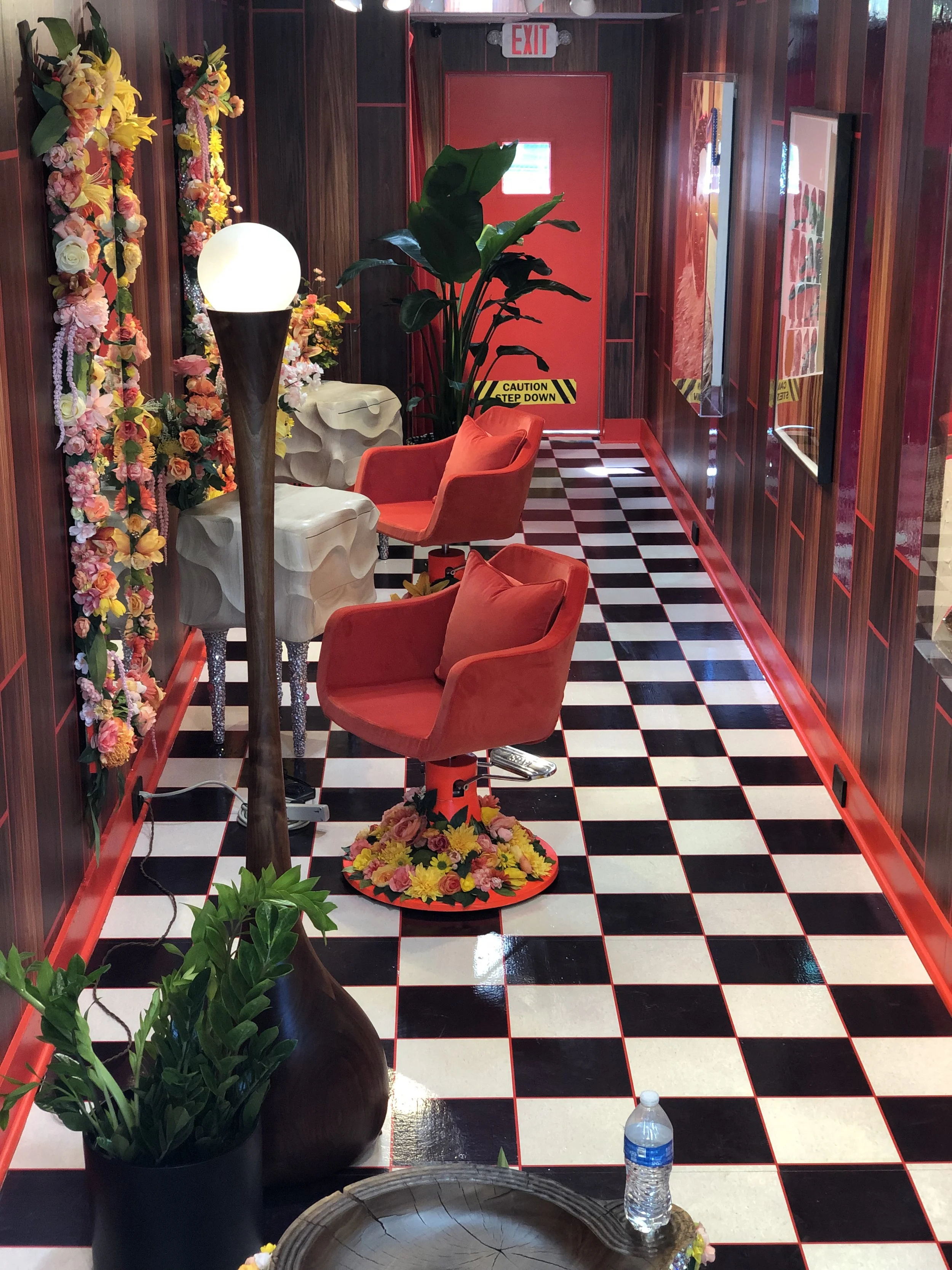 A colorful, retro-style room with red walls and black-and-white checkered flooring. There are red chairs with flowers around the base, a tall lamp with a globe light, and large green plants. Floral decorations adorn the walls, and a red door with an 