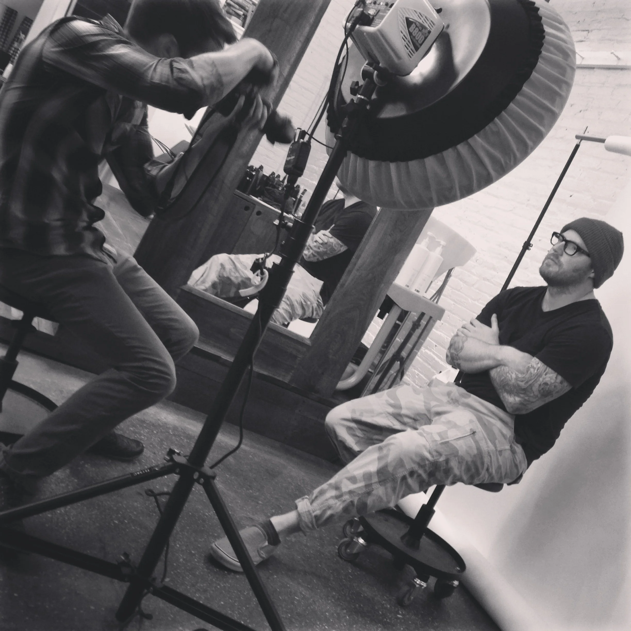 A man with a beanie and glasses sitting on a roller stool with crossed arms, facing a camera setup, while another person is filming him in a studio with a white brick wall and a mirror.